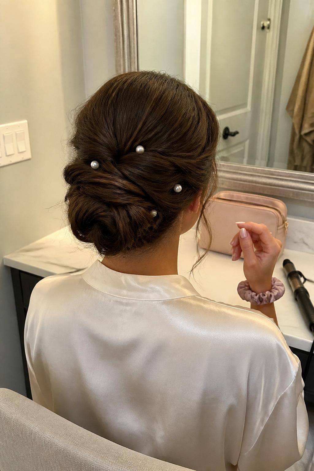 Image prompt: Photo of a woman wearing a soft twisted low chignon with overlapping side sections and small pearl pins, back-of-head view to highlight the details, bathroom vanity mirror with a makeup bag nearby, soft indoor lighting, containing a curling iron on the counter and a silk scrunchie around her wrist, realistic wedding guest getting-ready moment, latest iPhone photo quality.