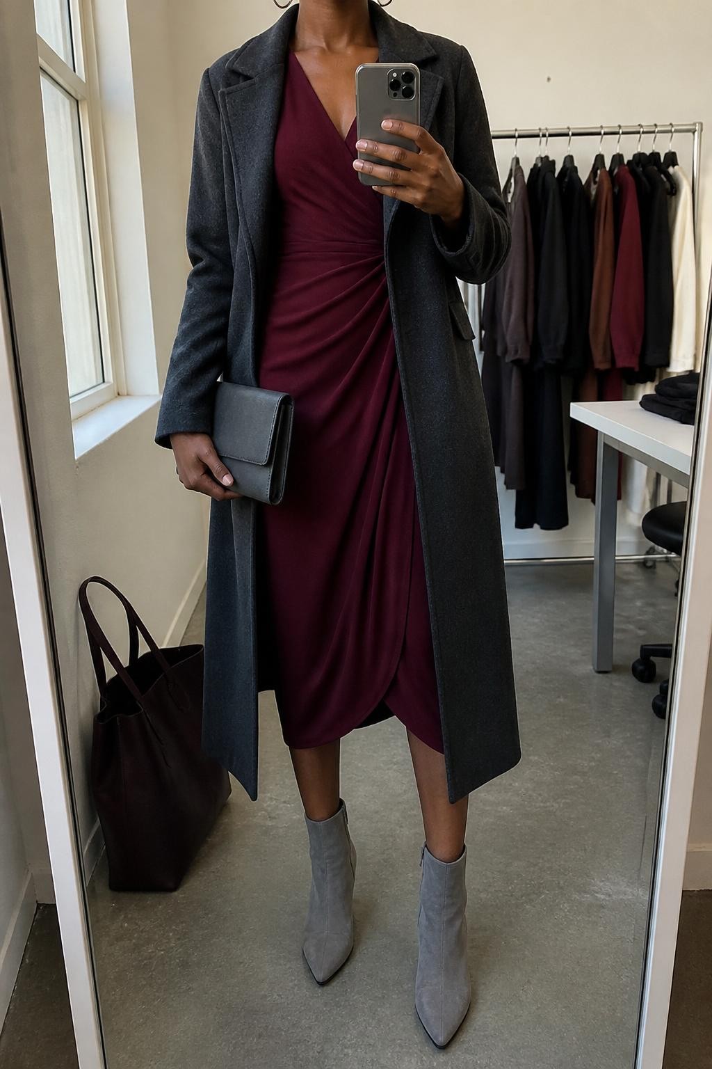 mirror selfie of a Black woman wearing the exact outfit described above, clearly showing gray suede ankle boots, a burgundy long-sleeve wrap midi dress, a charcoal long coat worn open, a gray clutch, and silver hoop earrings, holding a phone in front of a mirror, cropped at NECK so the face does not show, with a relaxed casual selfie pose, in a fashion studio mirror corner, natural window light, with a tote bag resting on the floor and clothing rack with garments behind, latest iPhone photo quality.