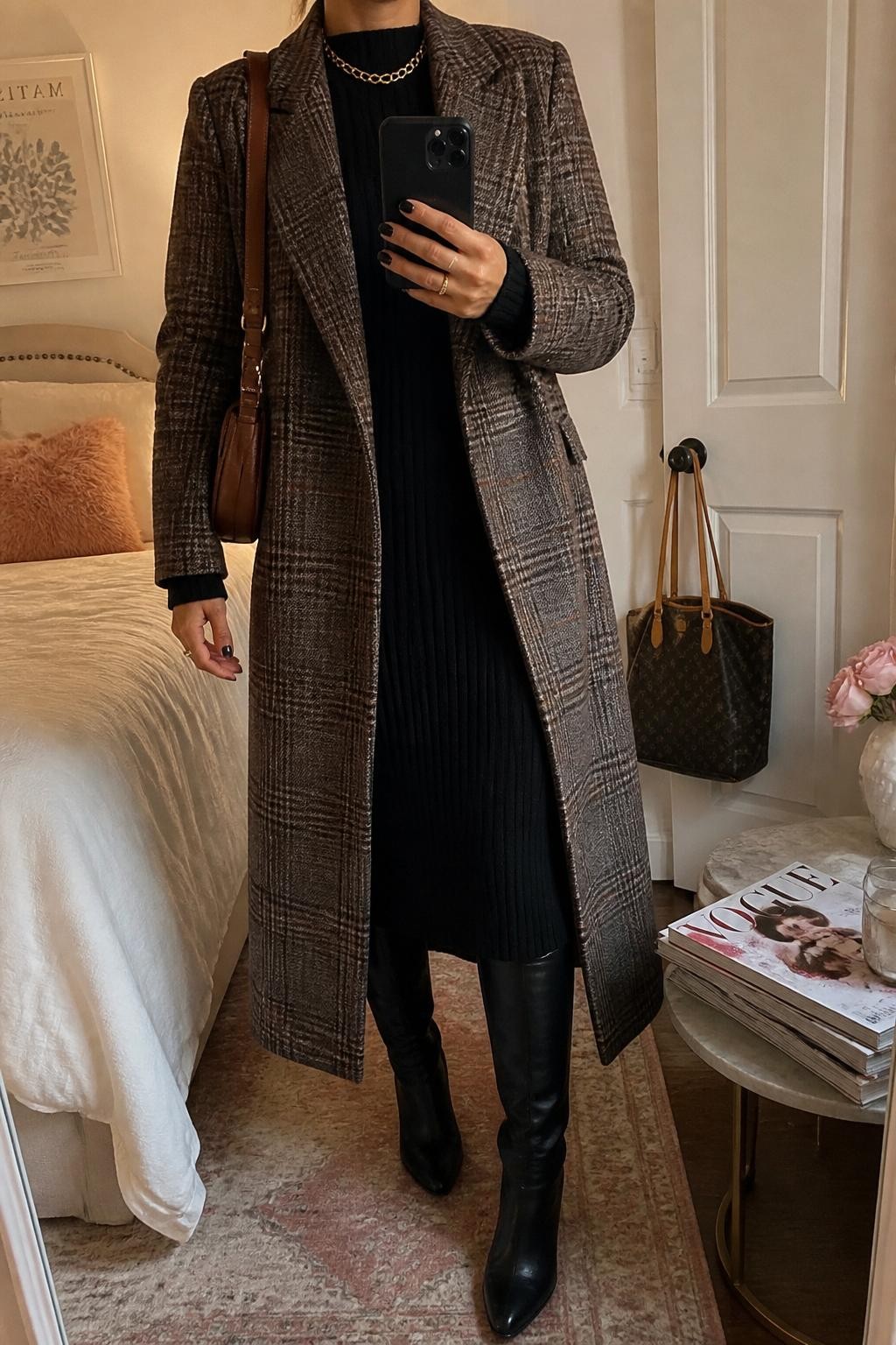 mirror selfie of a woman wearing the exact outfit described above, clearly showing a brown and gray plaid longline coat layered over a black ribbed knit midi dress, black knee-high boots, a brown leather shoulder bag, and gold chain necklace, holding a phone in front of a mirror, cropped at NECK so the face does not show, with a relaxed casual selfie pose, in a feminine-themed bedroom, warm moody lighting, with a handbag hanging on a door handle and a stack of fashion magazines on a side table, latest iPhone photo quality.
