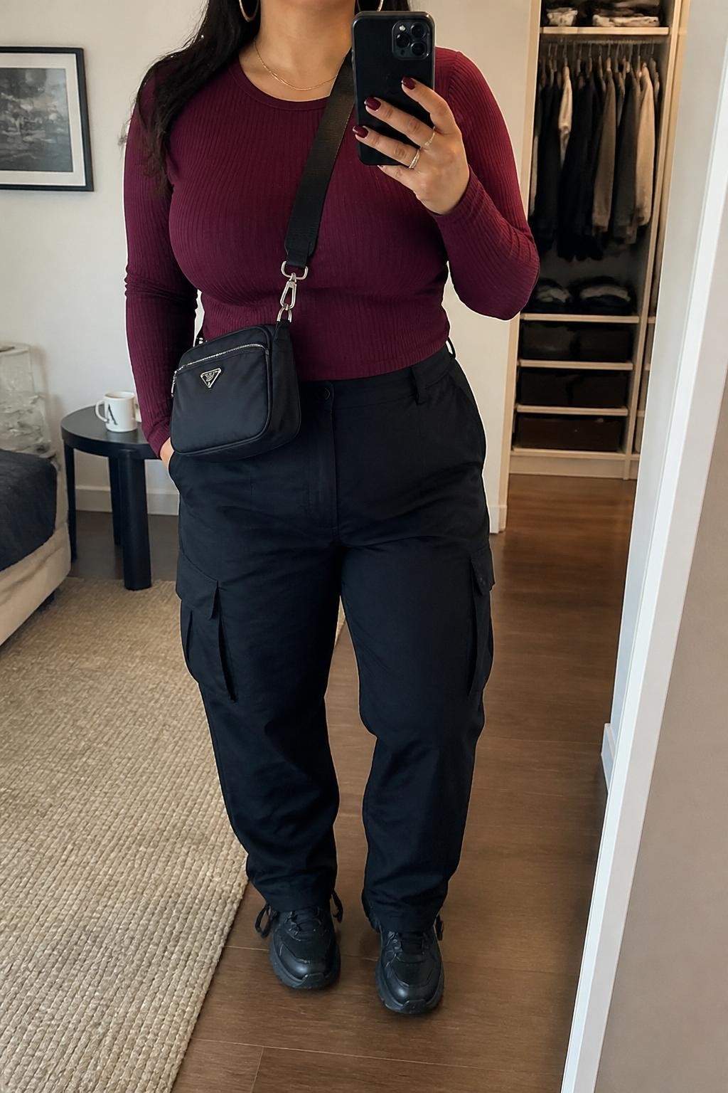 Mirror selfie of a Latina woman wearing a burgundy ribbed long-sleeve top, black high-waisted cargo pants, black chunky sneakers, gold hoop earrings, and a black nylon crossbody bag, holding a phone in front of a mirror, cropped at NECK so the face does not show, relaxed casual selfie pose, in a stylish city apartment, soft indoor lighting, with an open closet with clothes visible and a coffee mug on a side table, latest iPhone photo quality.