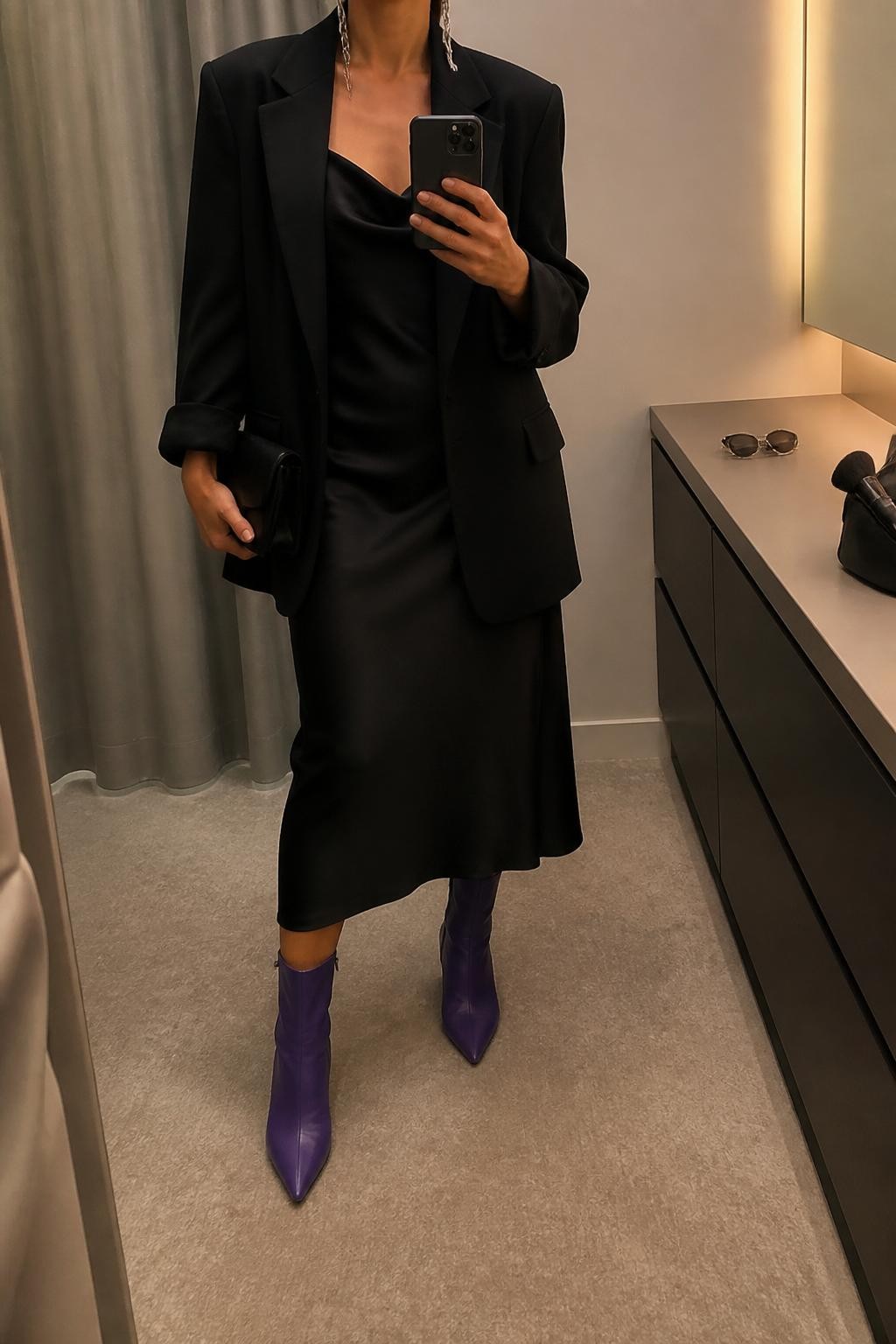 mirror selfie of a woman wearing the exact outfit described above, clearly showing violet pointed ankle boots, a black satin midi dress with a soft cowl neckline, a black oversized blazer, a small black clutch, and silver drop earrings, holding a phone in front of a mirror, cropped at NECK so the face does not show, with a relaxed casual selfie pose, in a minimal dressing room, warm moody lighting, with sunglasses placed on a dresser and a makeup brush or cosmetic bag on the counter, latest iPhone photo quality.
