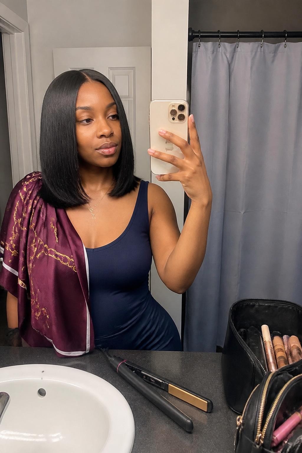 Image prompt: Photo of a Black woman wearing a smooth shoulder-length silk-press lob with softly bent ends and a clean middle part, candid mirror selfie, bathroom vanity mirror with a makeup bag nearby, soft indoor lighting, containing a flat iron on the counter and a satin scarf draped over her shoulder, realistic getting-ready moment, latest iPhone photo quality.