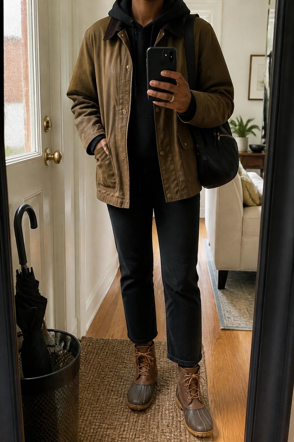 mirror selfie of a Black woman wearing the exact outfit described above, clearly showing a tan waxed jacket layered over a black hoodie, black cropped straight-leg jeans, brown waterproof lace-up boots, a black nylon backpack purse, and small silver hoop earrings, holding a phone in front of a mirror, cropped at NECK so the face does not show, with a relaxed casual selfie pose, in a cozy apartment entryway mirror, natural rainy daylight, with a wet umbrella near the door and boots on a mat, latest iPhone photo quality.