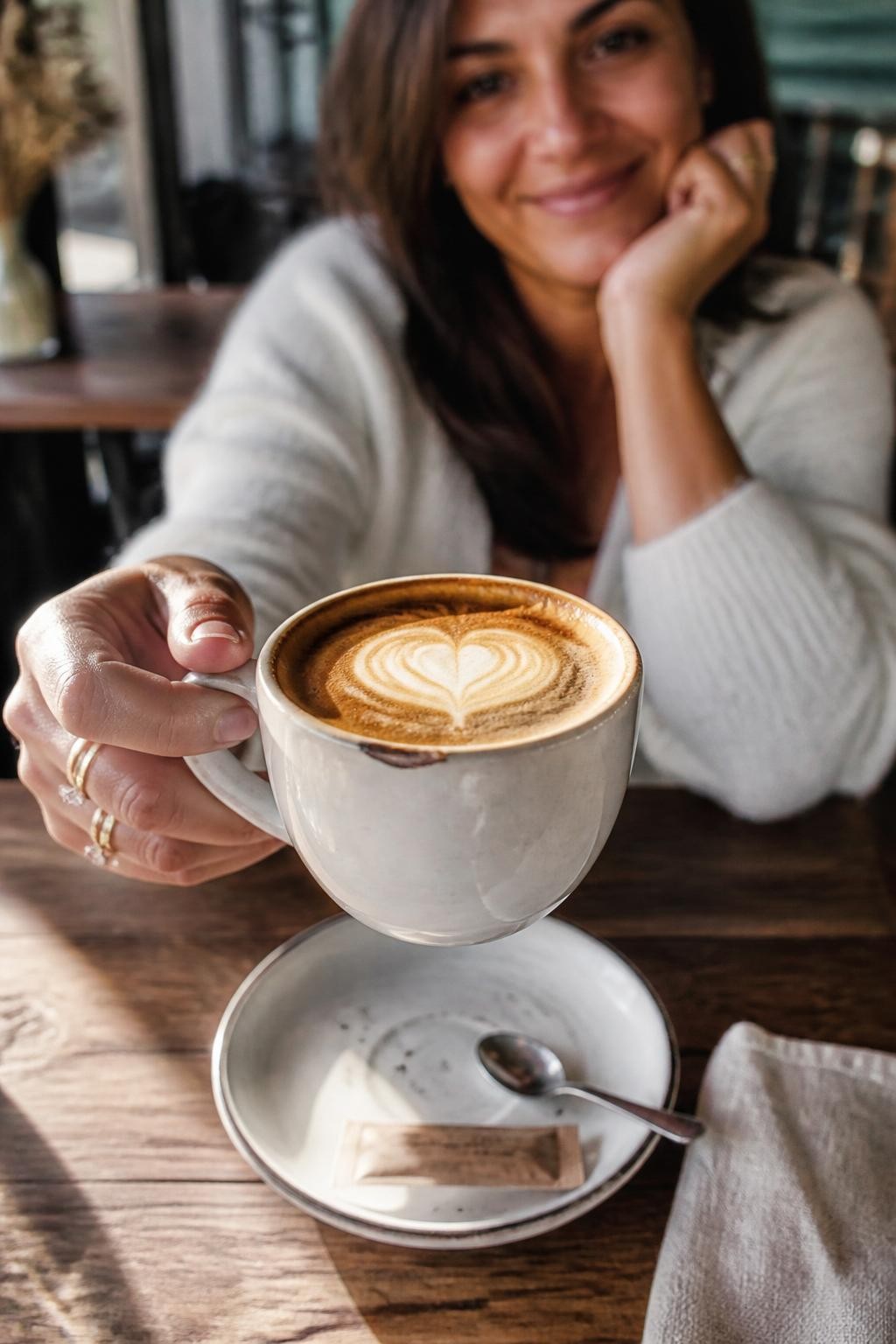 A realistic close-up cafe photo of an adult woman with medium skin tone sitting at a wooden cafe table, wearing a soft white cardigan and delicate rings, holding a cappuccino cup toward the camera so the heart-shaped foam art is in sharp focus while her smiling face is softly blurred in the background, one elbow resting on the table, small spoon on the saucer, sugar packet, linen napkin, sunlight hitting the coffee surface, natural hand texture, slight chipped edge on the ceramic cup, cozy cafe details, DSLR shallow depth of field.