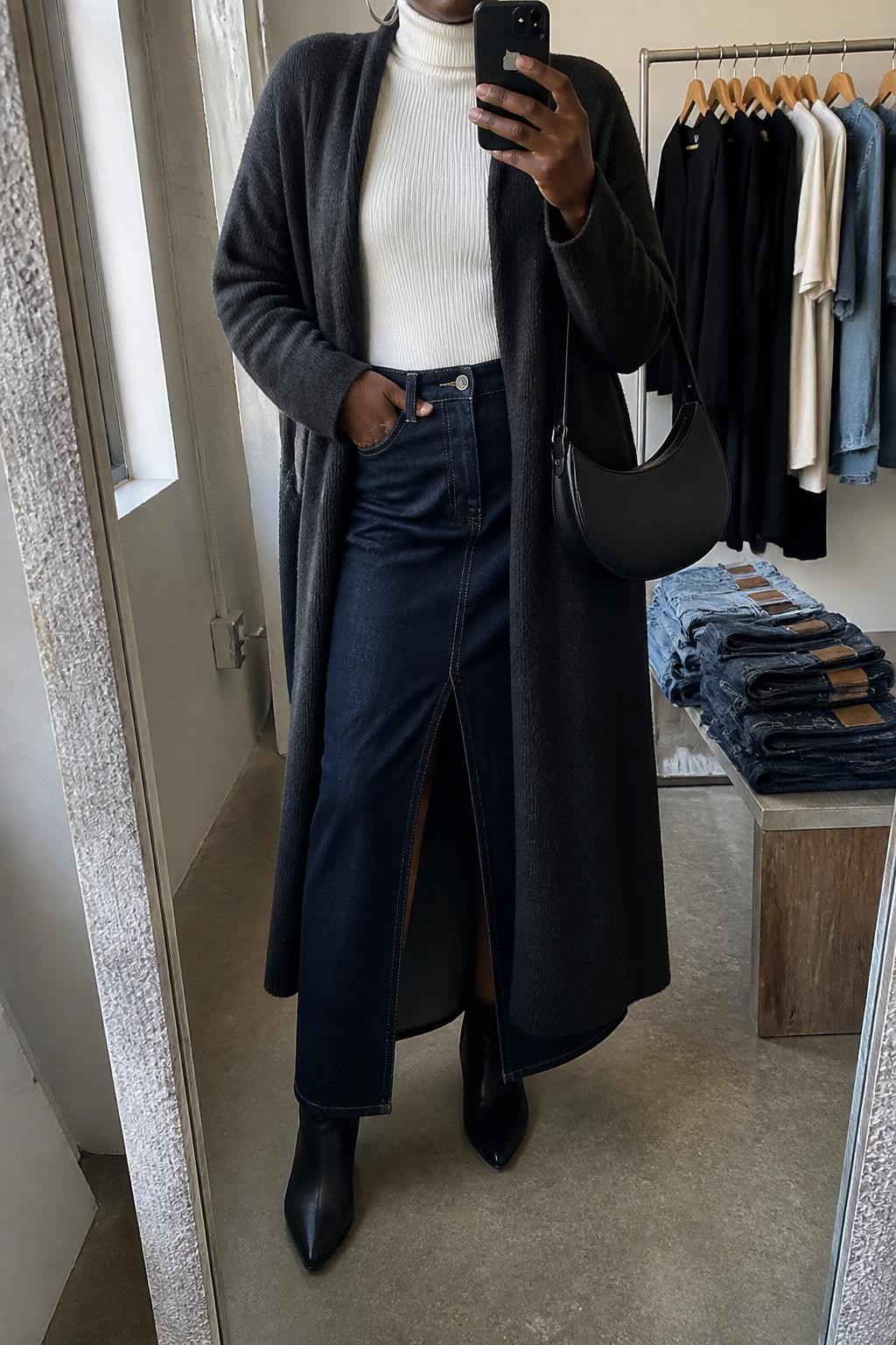 Mirror selfie of a Black woman wearing a charcoal long coatigan, white ribbed turtleneck, dark denim maxi skirt with front slit, black ankle boots, silver hoop earrings, and a black saddle bag, holding a phone in front of a mirror, cropped at NECK so the face does not show, relaxed casual selfie pose, in a fashion studio mirror corner, natural window light, with a clothing rack with garments behind and folded denim jeans on a bench, latest iPhone photo quality.