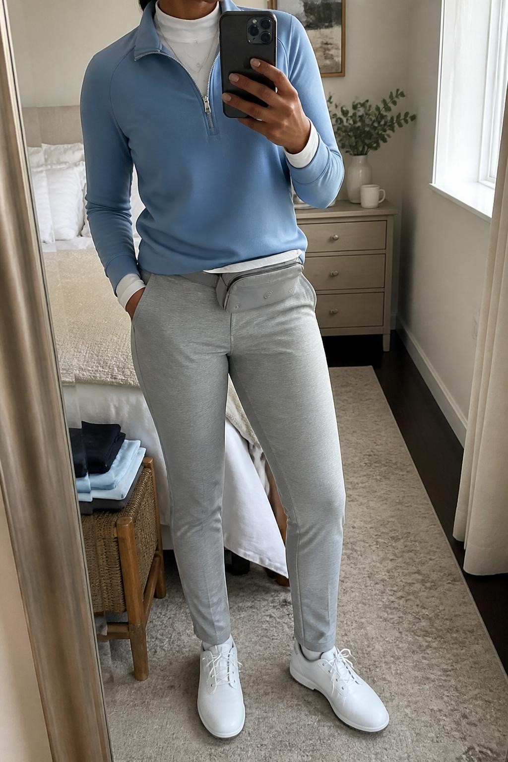 Mirror selfie of a Black woman wearing a dusty blue half-zip golf pullover over a white mock-neck performance top, paired with heather gray slim golf trousers, clearly showing white waterproof golf shoes, a dusty blue cap, and a light gray belt bag, holding an iPhone in front of a cozy bedroom mirror setup, cropped at the neck so the face does not show, relaxed casual selfie pose, natural window light, folded clothes on a bench and a coffee mug on a bedside table, natural unstaged setting, latest iPhone photo quality.