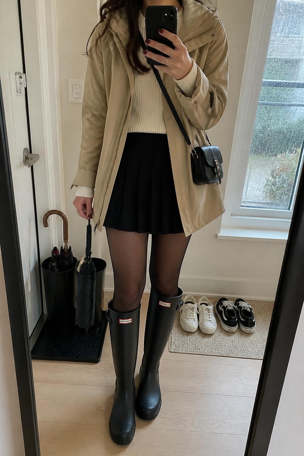 mirror selfie of a woman wearing the exact outfit described above, clearly showing a beige hooded raincoat layered over a cream ribbed sweater, black pleated mini skirt, sheer black tights, black knee-high rain boots, a small black crossbody bag, and a compact black umbrella in hand, holding a phone in front of a mirror, cropped at NECK so the face does not show, with a relaxed casual selfie pose, in a cozy apartment entryway mirror, soft rainy window light, with a wet umbrella stand near the door and shoes on a mat, latest iPhone photo quality.