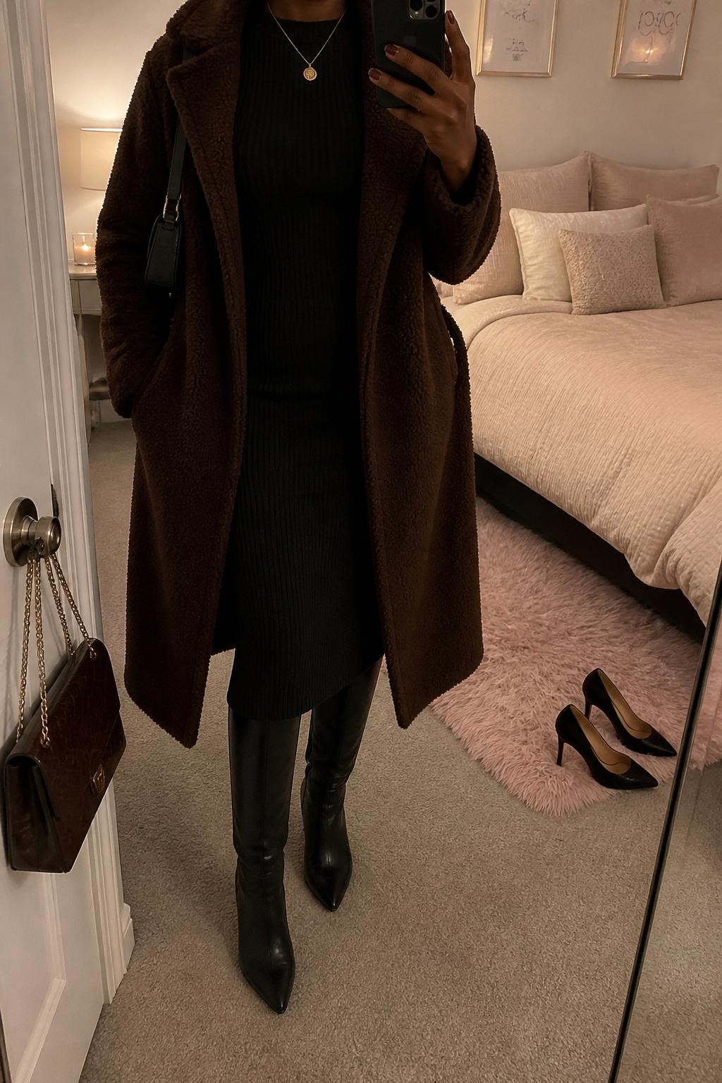 Mirror selfie of a Black woman wearing a chocolate brown teddy coat, black ribbed long-sleeve midi dress, black knee-high boots, gold pendant necklace, and a small brown shoulder bag, holding a phone in front of a mirror, cropped at NECK so the face does not show, relaxed casual selfie pose, in a feminine-themed bedroom, warm moody lighting, with a pair of heels on the floor nearby and a handbag hanging on a door handle, latest iPhone photo quality.