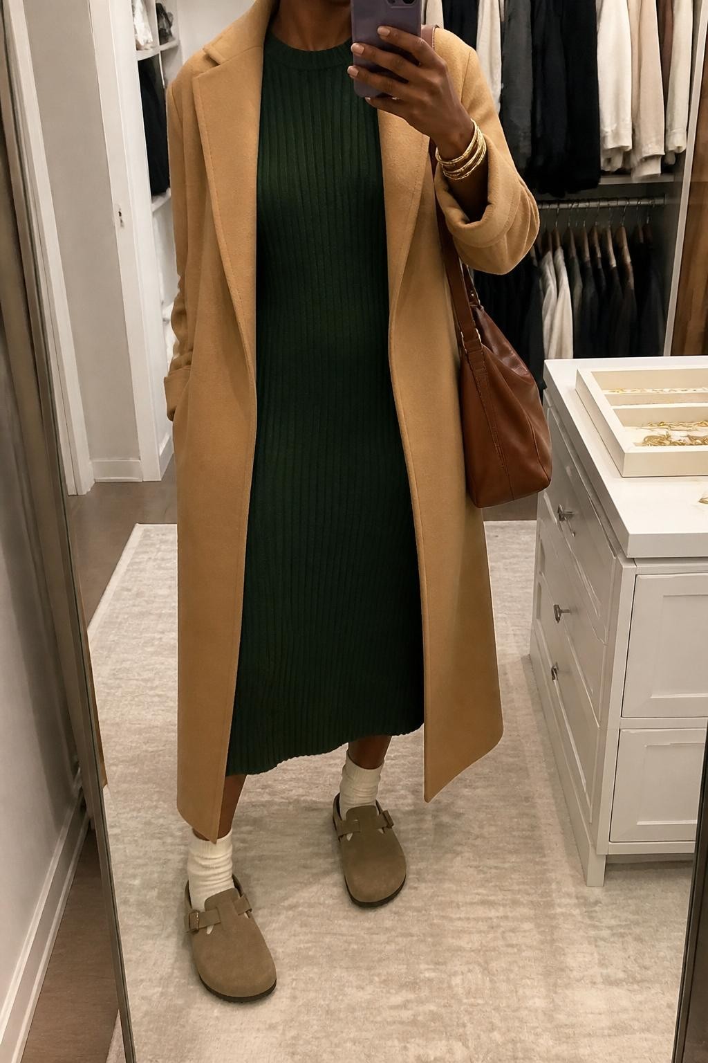 mirror selfie of a Black woman wearing the exact outfit described above, clearly showing mink taupe Birkenstock-style clogs with oatmeal socks, a forest green ribbed sweater midi dress, a camel long coat worn open, a brown leather shoulder bag, and gold bangles, holding a phone in front of a mirror, cropped at NECK so the face does not show, with a relaxed casual selfie pose, in a walk-in closet mirror, soft indoor lighting, with an open closet with clothes visible and a jewelry tray on the vanity, latest iPhone photo quality.