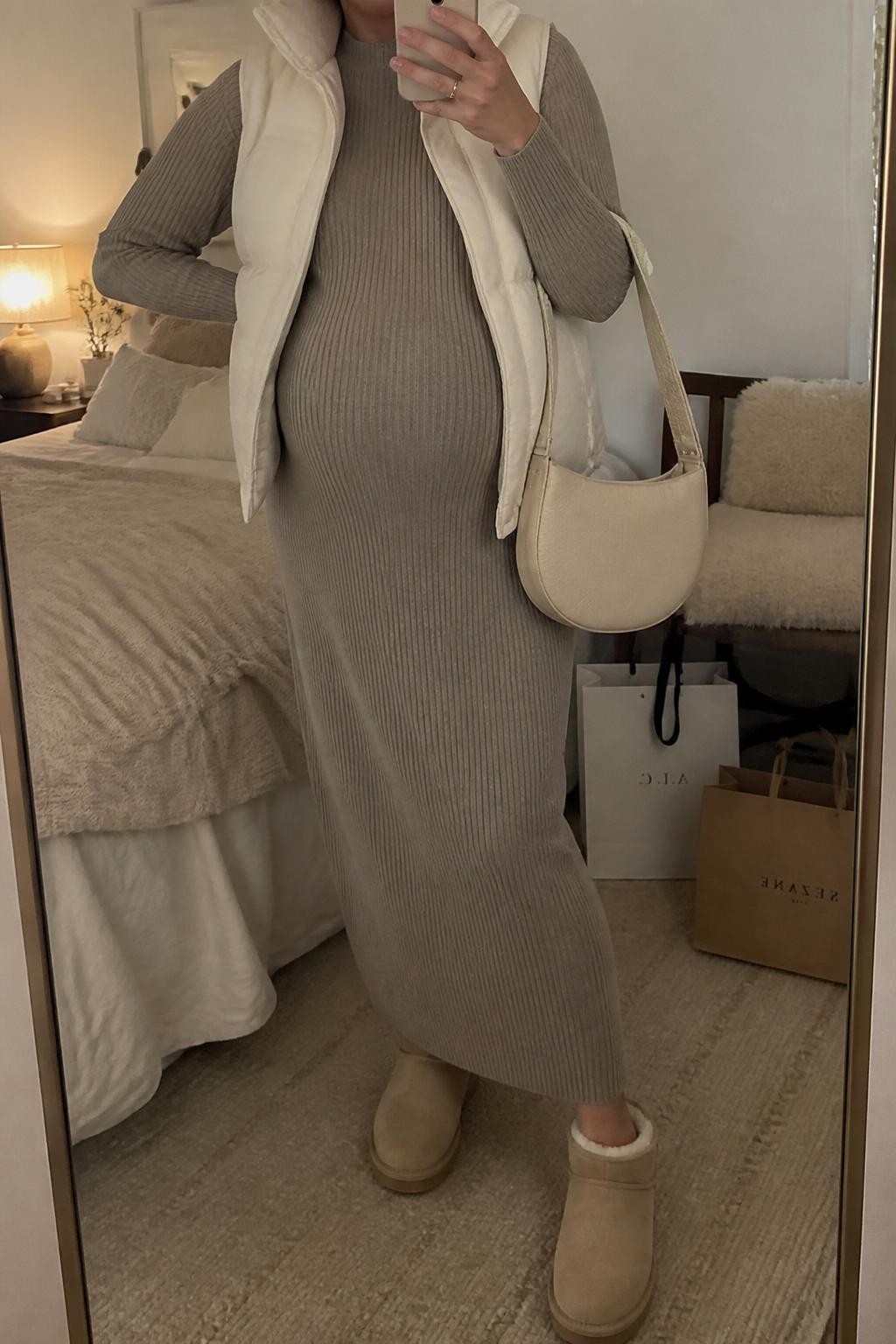 mirror selfie of a pregnant woman wearing a taupe long sleeve knit maternity maxi dress, cream quilted puffer vest, beige shearling-lined ankle boots, a cream knit beanie, and a beige crescent bag, clearly showing the same clothing items, colors, fabrics, and accessories listed in the Outfit Pieces section, holding a phone in front of a mirror, cropped at NECK so the face does not show, with a relaxed casual selfie pose, in a cozy bedroom mirror setup, warm moody lighting, including shopping bags beside a chair and soft pillows on an unmade bed, latest iPhone photo quality.