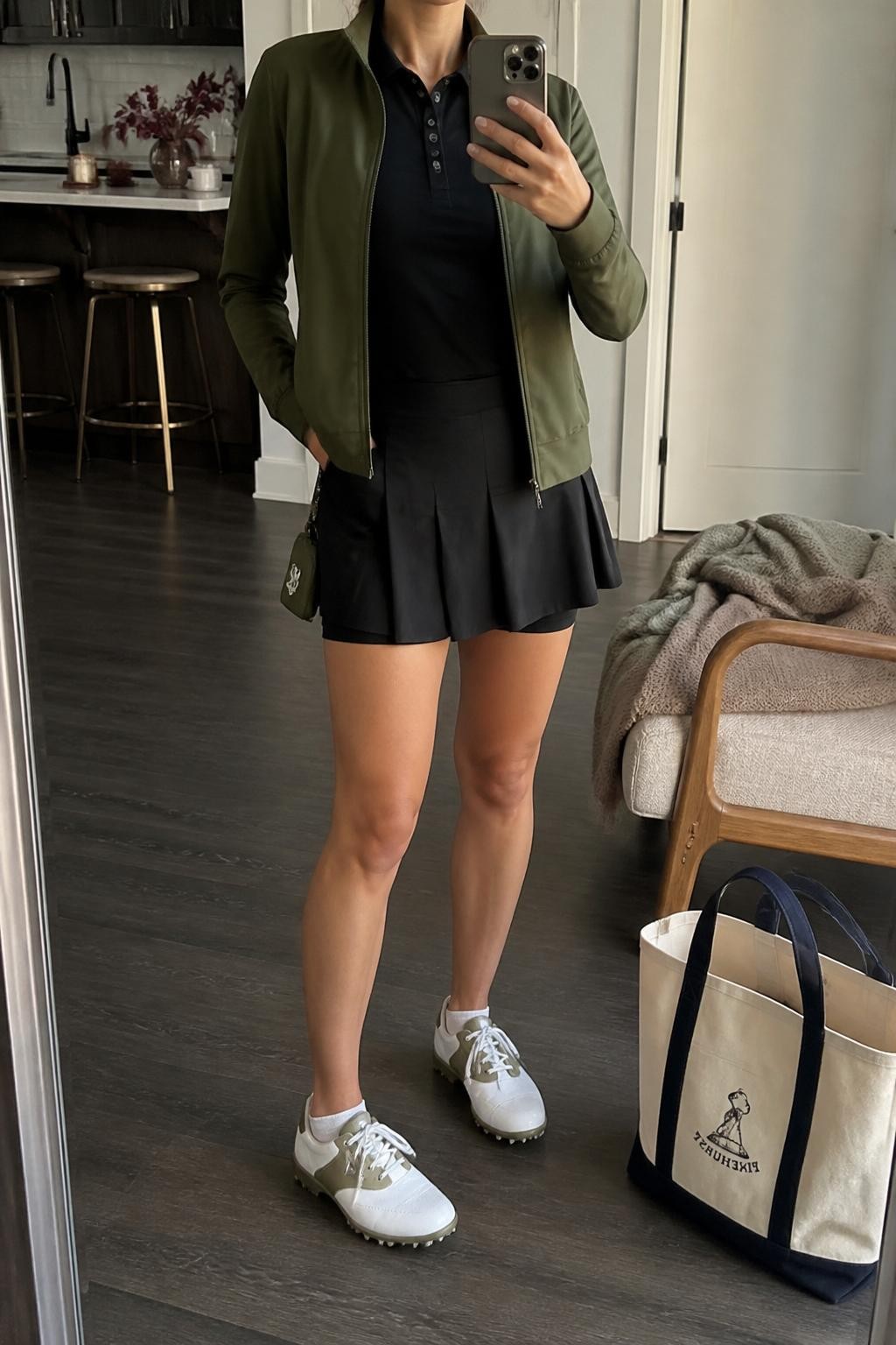 Mirror selfie of a woman wearing an olive green lightweight zip-front golf jacket over a fitted black collared polo, paired with a black pleated golf skort with built-in shorts, clearly showing white-and-olive golf shoes, a black cap, and a small olive golf pouch, holding an iPhone in front of a stylish city apartment mirror, cropped at the neck so the face does not show, relaxed casual selfie pose, soft indoor lighting, a tote bag resting on the floor and a folded blanket on a chair, natural unstaged setting, latest iPhone photo quality.