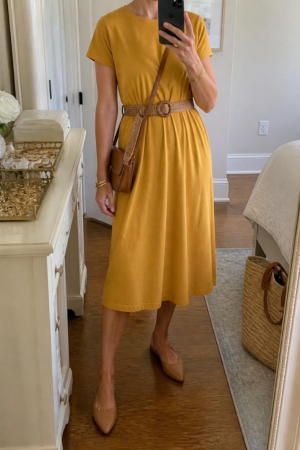mirror selfie of a woman wearing the exact outfit described above, clearly showing a mustard short-sleeve cotton midi dress, tan slingback flats, a caramel woven belt, a small brown crossbody bag, and gold bangles, holding a phone in front of a mirror, cropped at NECK so the face does not show, with a relaxed casual selfie pose, in a feminine bedroom mirror setup, natural daylight, with a jewelry tray on the vanity and a straw tote resting near the wall, latest iPhone photo quality.