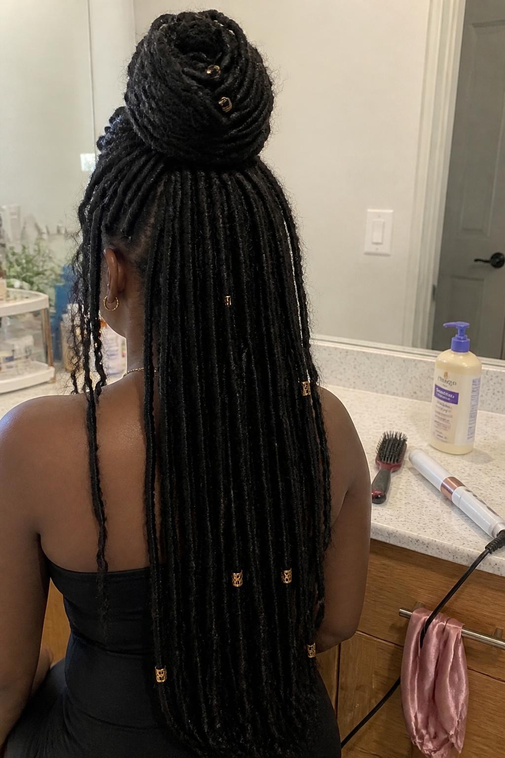 Image prompt: Photo of a Black woman wearing long soft locs styled into a large half-up bun with a few gold cuffs along the loose lengths, back-of-head view to highlight the details, bathroom vanity mirror with a hairbrush and lotion nearby, soft indoor lighting, containing a curling iron on the counter and a satin scarf hanging from a drawer handle, realistic getting-ready moment, latest iPhone photo quality.