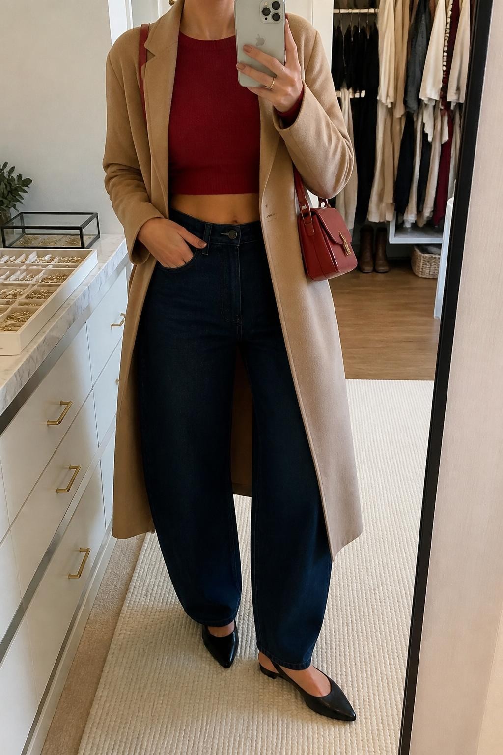 mirror selfie of a woman wearing the exact outfit described above, clearly showing dark indigo barrel jeans, a red cropped knit sweater, black slingback flats, a camel long coat worn open, a small red shoulder bag, and gold stud earrings, holding a phone in front of a mirror, cropped at NECK so the face does not show, with a relaxed casual selfie pose, in a walk-in closet mirror, soft indoor lighting, with an open closet with clothes visible and a jewelry tray on the vanity, latest iPhone photo quality.