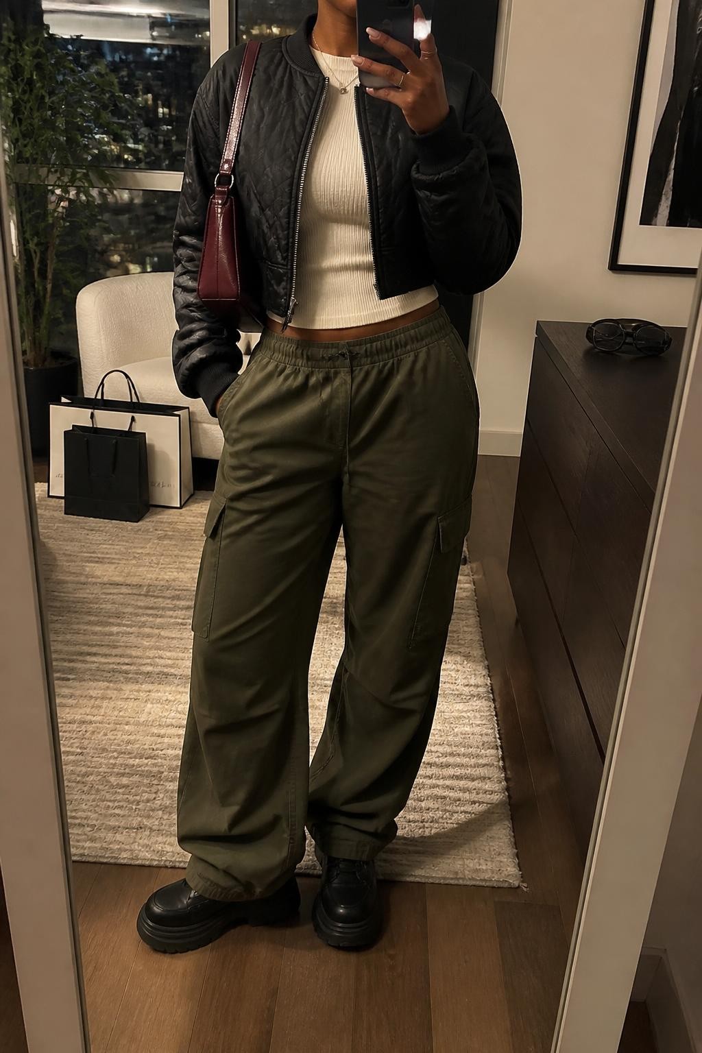 Mirror selfie of a Black woman wearing a cropped black quilted bomber jacket, fitted cream ribbed crew-neck top, loose olive-green cargo pants, chunky black leather sneakers, and a burgundy nylon shoulder bag, clearly showing the exact clothing items, colors, fabrics, and accessories listed, holding a phone in front of a stylish city apartment mirror, cropped at the neck so the face does not show, relaxed casual selfie pose, warm moody lighting, shopping bags beside a chair and sunglasses placed on a dresser, realistic city setting, latest iPhone photo quality.