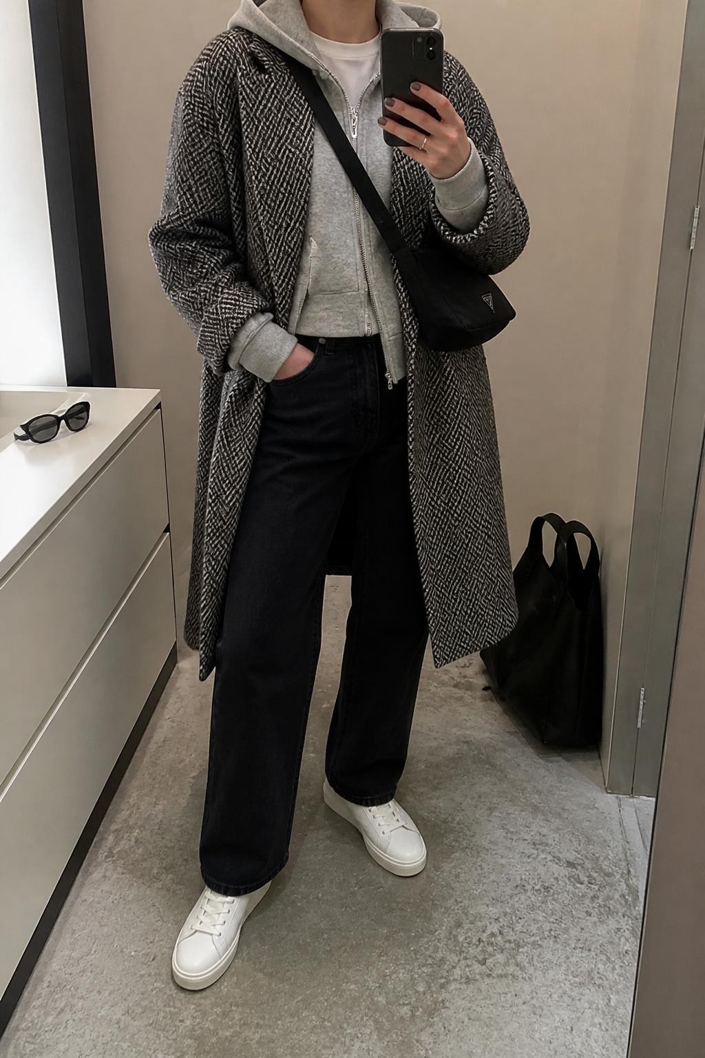 Mirror selfie of a woman wearing a black-and-gray herringbone long coat over a light gray fitted hoodie and a white crew-neck T-shirt, clearly showing black straight-leg jeans, white leather sneakers, and a black nylon crossbody bag, holding an iPhone in front of a minimal dressing room mirror, cropped at the neck so the face does not show, relaxed casual selfie pose, soft indoor lighting, a tote bag resting on the floor and sunglasses placed on a dresser, natural unstaged setting, latest iPhone photo quality.