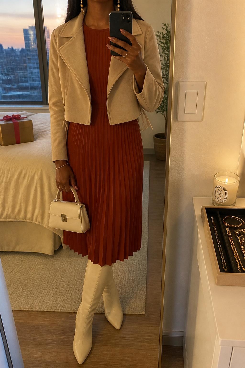 mirror selfie of a Black woman wearing the exact outfit described above, clearly showing a rust pleated long-sleeve midi dress, cream knee-high boots, a beige cropped jacket, a cream mini handbag, and gold drop earrings, holding a phone in front of a mirror, cropped at NECK so the face does not show, with a relaxed casual selfie pose, in a stylish apartment mirror setup, evening golden hour, with a wrapped gift box on the bed and a jewelry tray on the vanity, latest iPhone photo quality.