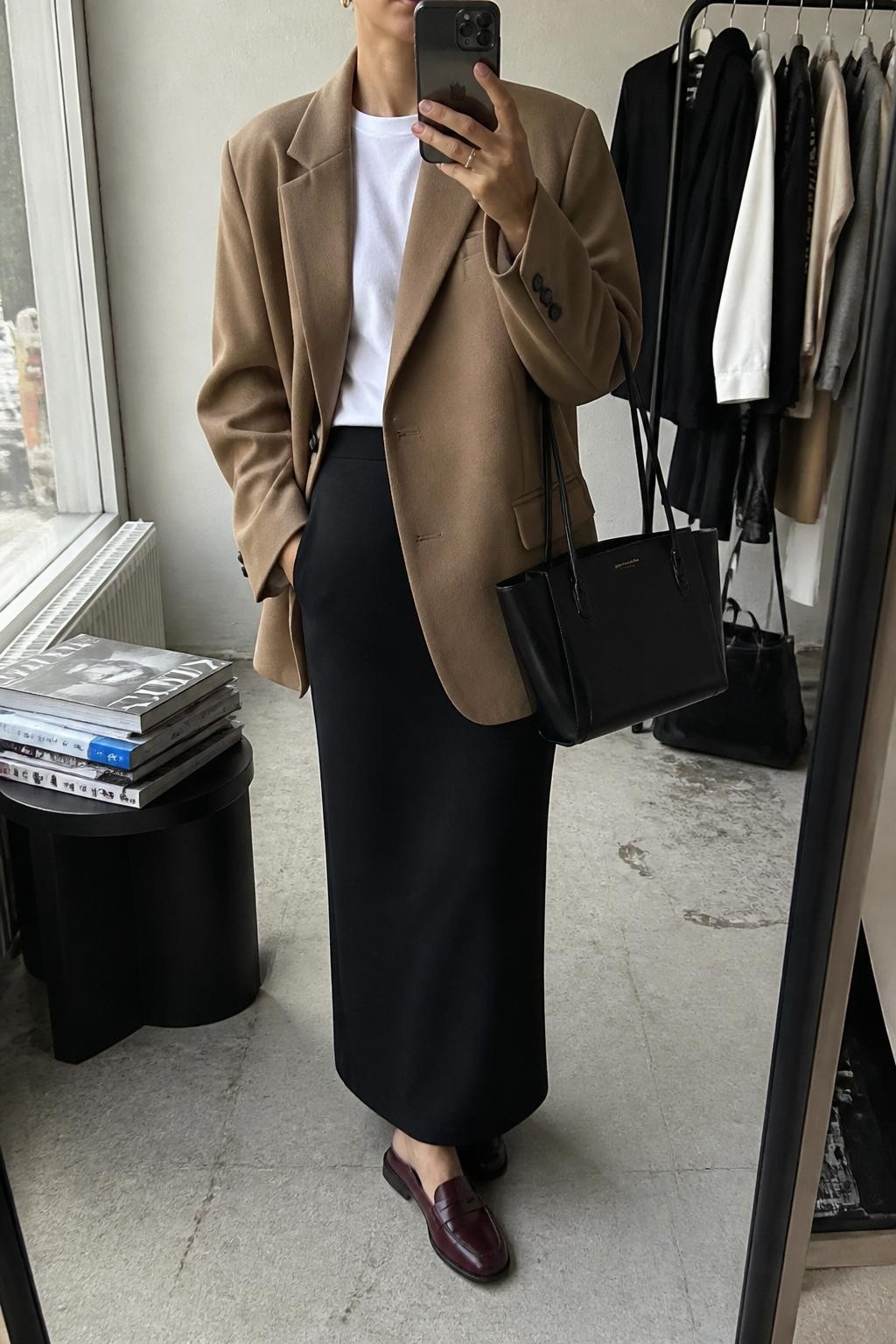 Mirror selfie of a woman wearing a camel oversized wool blazer over a fitted white crew-neck T-shirt and a black ankle-length column skirt, clearly showing burgundy leather loafers, a structured black tote, and small gold stud earrings, holding an iPhone in front of a fashion studio mirror corner, cropped at the neck so the face does not show, relaxed casual selfie pose, natural window light, a stack of fashion magazines on a side table and a clothing rack with garments behind, natural unstaged setting, latest iPhone photo quality.