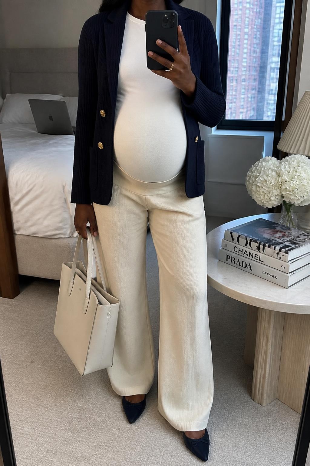 mirror selfie of a pregnant Black woman wearing a navy knit blazer, white fitted maternity tee, cream maternity wide-leg trousers, navy pointed flats, and a cream structured tote bag, clearly showing the same clothing items, colors, fabrics, and accessories listed in the Outfit Pieces section, holding a phone in front of a mirror, cropped at NECK so the face does not show, with a relaxed casual selfie pose, in a stylish city apartment, soft indoor lighting, including stack of fashion magazines on a side table and laptop on the bed, latest iPhone photo quality.