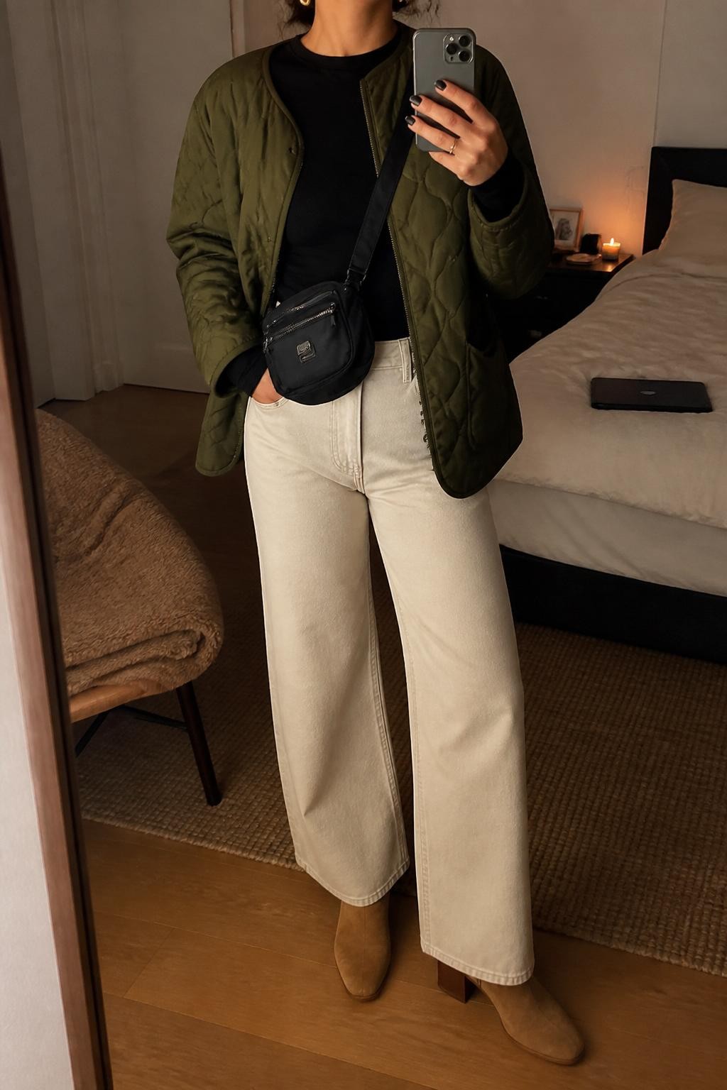 mirror selfie of a woman wearing the exact outfit described above, clearly showing cream wide-leg jeans, a fitted black long-sleeve top, an olive quilted jacket, tan suede ankle boots, a black nylon crossbody bag, and small gold earrings, holding a phone in front of a mirror, cropped at NECK so the face does not show, with a relaxed casual selfie pose, in a cozy bedroom mirror setup, warm moody lighting, with a folded blanket on a chair and a laptop or tablet on the bed, latest iPhone photo quality.
