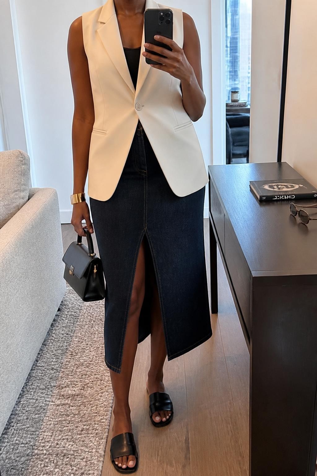 mirror selfie of a Black woman wearing the exact outfit described above, clearly showing a dark denim midi skirt with a front slit, cream sleeveless blazer worn buttoned over a fitted black tank, black leather slides, a structured black mini bag, and gold cuff bracelet, holding a phone in front of a mirror, cropped at NECK so the face does not show, with a relaxed casual selfie pose, in a stylish apartment mirror, soft indoor lighting, with a book on a side table and sunglasses placed on a dresser, latest iPhone photo quality.