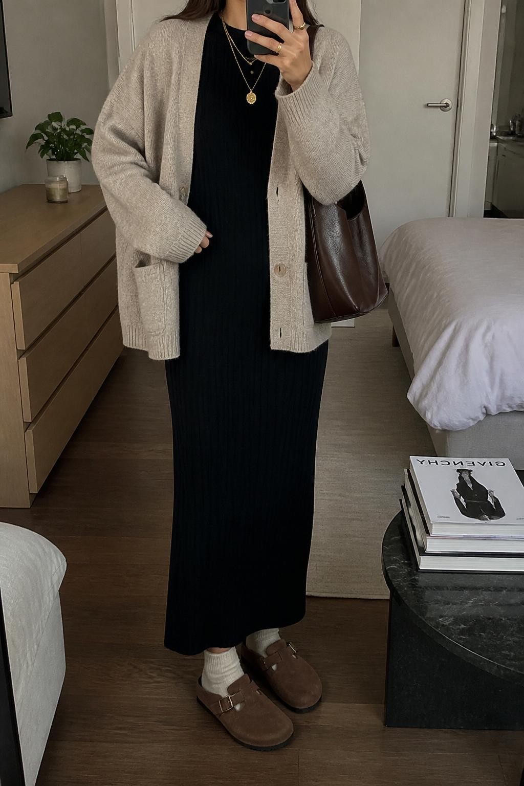 mirror selfie of a woman wearing the exact outfit described above, clearly showing tobacco brown suede Birkenstock-style clogs with cream socks, a black long-sleeve ribbed maxi dress, an oversized oatmeal cardigan, a brown leather shoulder bag, and gold layered necklaces, holding a phone in front of a mirror, cropped at NECK so the face does not show, with a relaxed casual selfie pose, in a stylish city apartment, soft indoor lighting, with a small plant on a dresser and a stack of fashion magazines on a side table, latest iPhone photo quality.