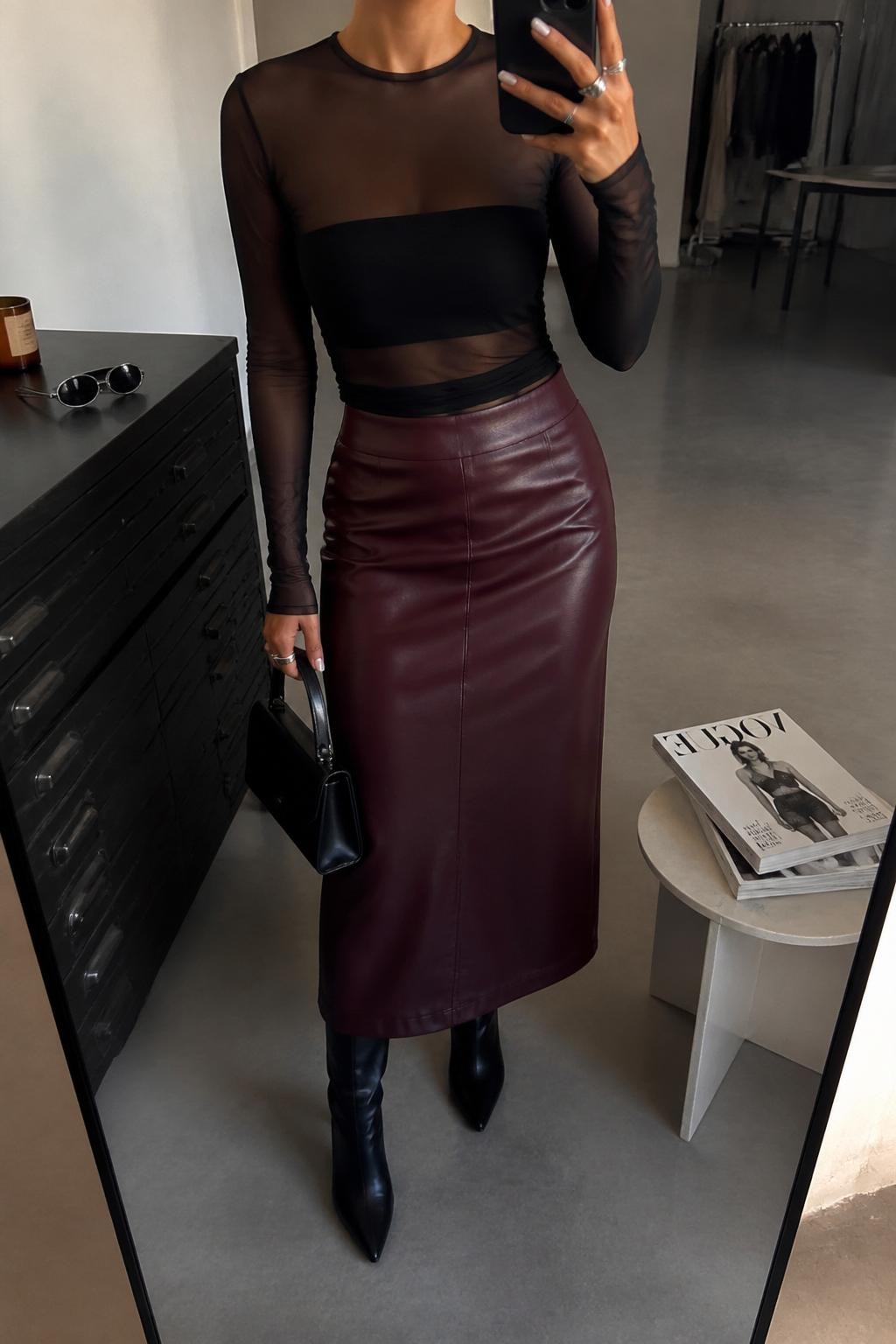 mirror selfie of a woman wearing a black sheer mesh long-sleeve top layered over a black bandeau, oxblood faux leather midi skirt, black pointed heeled boots, silver oval sunglasses, and a black structured mini bag, clearly showing the same clothing items, colors, fabrics, and accessories listed in the Outfit Pieces section, holding a phone in front of a mirror, cropped at NECK so the face does not show, with a relaxed casual selfie pose, in a fashion studio mirror corner, warm moody lighting, including sunglasses placed on a dresser and a stack of fashion magazines on a side table, latest iPhone photo quality.