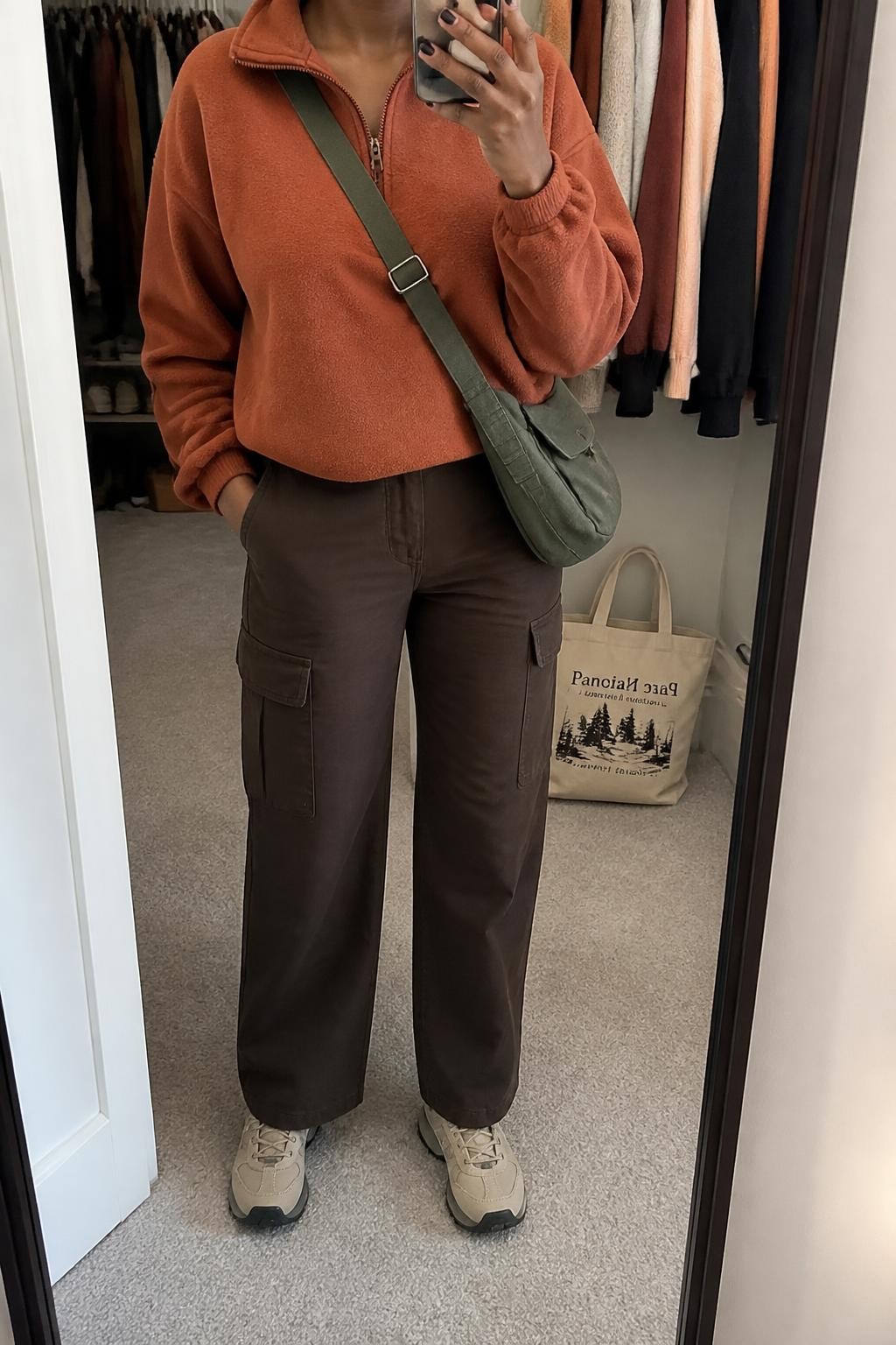 Mirror selfie of a Black woman wearing a rust-orange half-zip fleece pullover, chocolate-brown straight-leg cargo pants, beige waterproof hiking sneakers, and a dark green canvas crossbody bag, clearly showing the exact clothing items, colors, fabrics, and accessories listed, holding a phone in front of a walk-in closet mirror, cropped at the neck so the face does not show, relaxed casual selfie pose, soft indoor lighting, an open closet with clothes visible and a tote bag resting on the floor, realistic and uncluttered, latest iPhone photo quality.