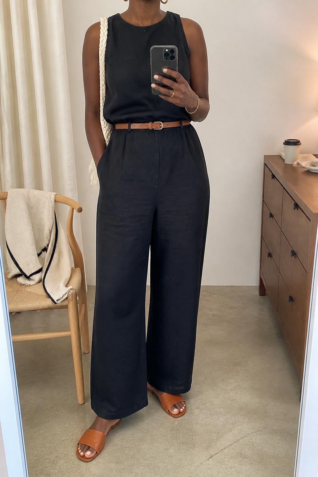 mirror selfie of a Black woman wearing the exact outfit described above, clearly showing a black sleeveless linen jumpsuit with a relaxed wide-leg cut, tan leather slide sandals, a cream woven shoulder bag, gold hoop earrings, and a slim brown belt, holding a phone in front of a mirror, cropped at NECK so the face does not show, with a relaxed casual selfie pose, in a minimal dressing room, warm natural light, with a coffee cup on the dresser and a folded cardigan on a chair, latest iPhone photo quality.