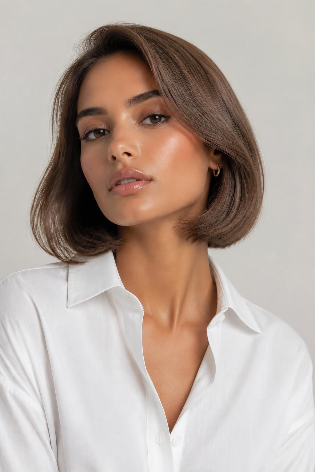 Image Prompt: A pretty woman with a chin-length light milk brown bob, smooth rounded bob shape with a soft bend at the ends, light creamy brown hair color with beige shine, medium skin tone, fresh natural makeup, glossy nude lips, small gold earrings, white button-up shirt, bright clean studio background, soft flattering lighting, realistic beauty editorial photography, detailed bob shape and soft brunette shine, no text, (no watermarks on images)
