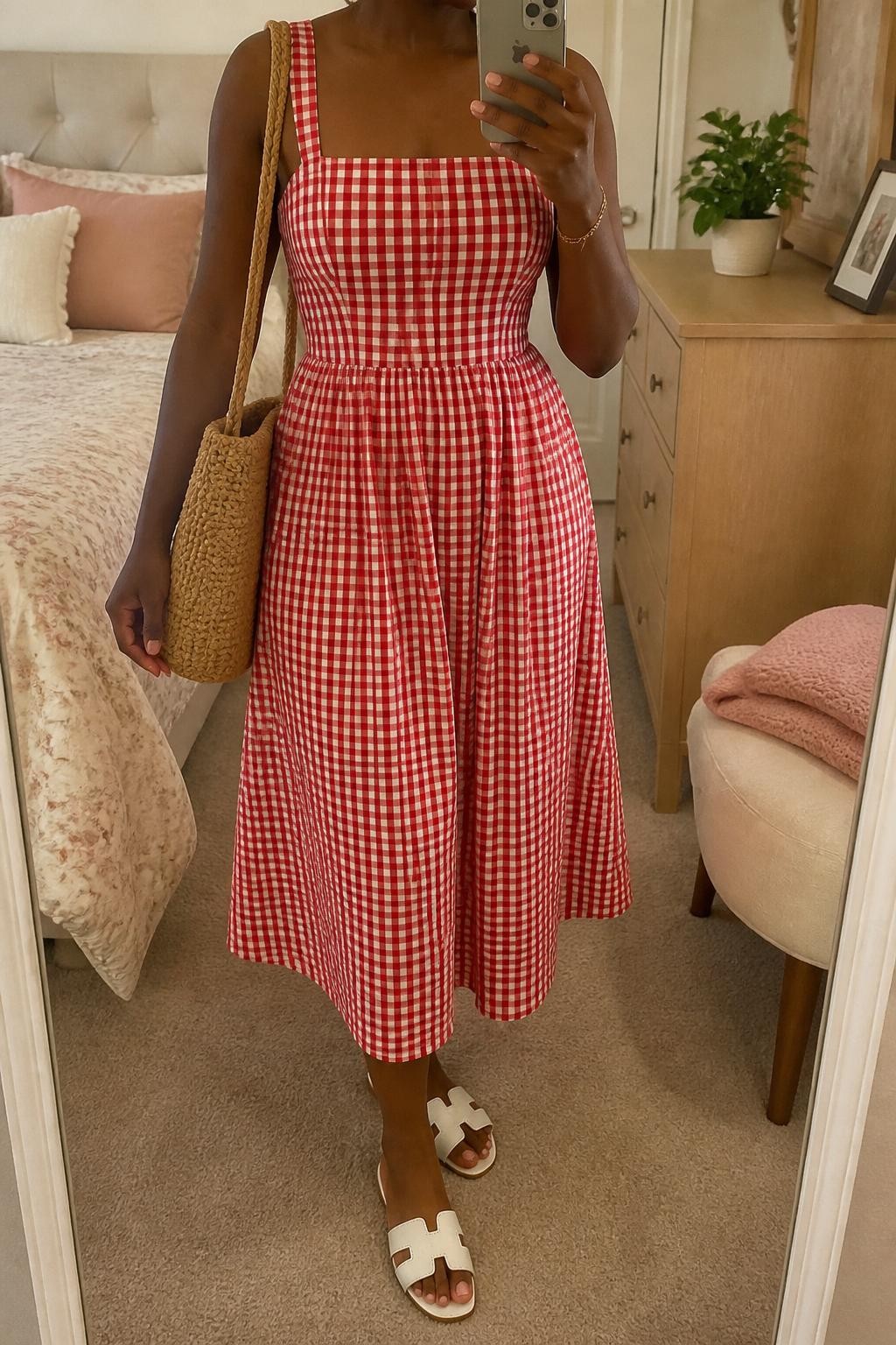 mirror selfie of a Black woman wearing a red and white gingham cotton midi dress with a fitted waist and softly flared skirt, white flat sandals, woven straw shoulder bag, pearl stud earrings, and a delicate gold bracelet, clearly showing the same clothing items, colors, fabrics, and accessories listed in the Outfit Pieces section, holding a phone in front of a mirror, cropped at NECK so the face does not show, with a relaxed casual selfie pose, in a feminine-themed bedroom, soft indoor lighting, including a small plant on a dresser and a folded blanket on a chair, latest iPhone photo quality.
