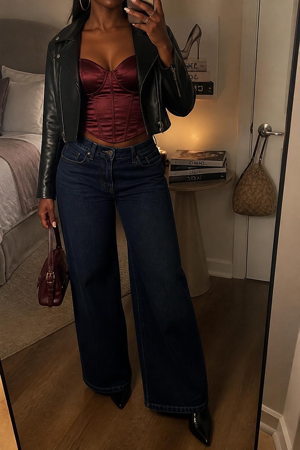 mirror selfie of a Black woman wearing the exact outfit described above, clearly showing a burgundy satin corset top, dark blue wide-leg jeans, black pointed heeled boots, a black cropped leather jacket, a burgundy mini handbag, and gold hoop earrings, holding a phone in front of a mirror, cropped at NECK so the face does not show, with a relaxed casual selfie pose, in a feminine-themed bedroom, warm moody lighting, with a handbag hanging on a door handle and a stack of fashion magazines on a side table, latest iPhone photo quality.