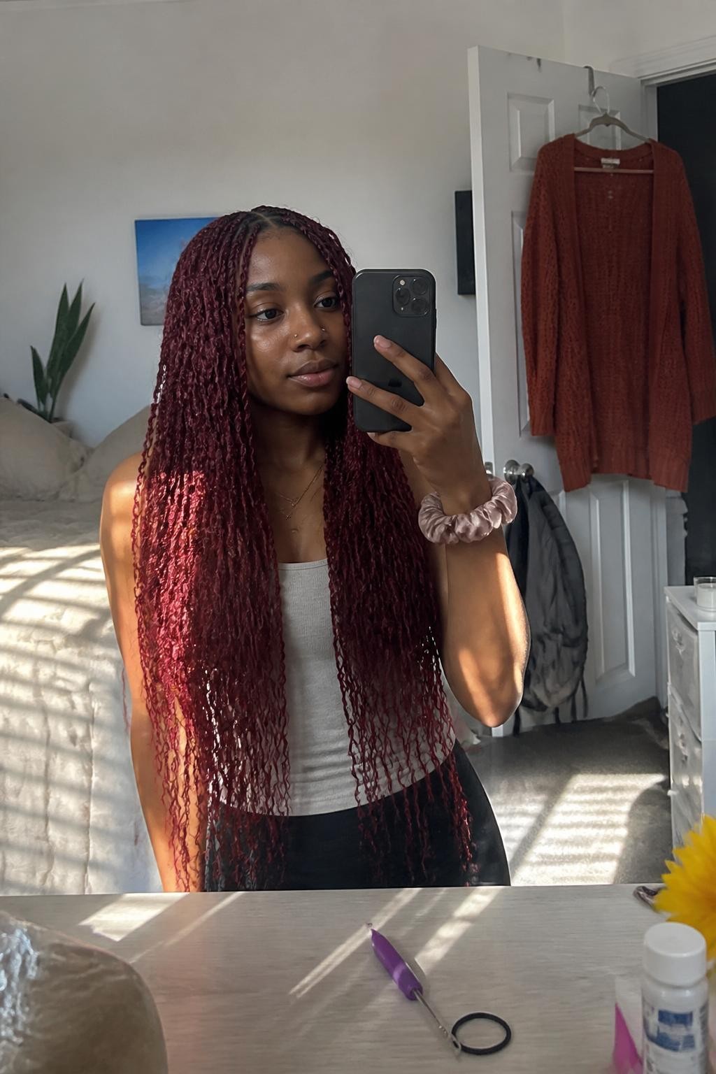Image prompt: Photo of a Black woman wearing long burgundy passion twists with soft loose texture and a clean middle part, candid mirror selfie, sun-drenched bedroom with a rust-colored cardigan hanging from an open closet door, natural window light, containing a crochet needle on the dresser and a silk scrunchie around her wrist, realistic home styling moment, latest iPhone photo quality.