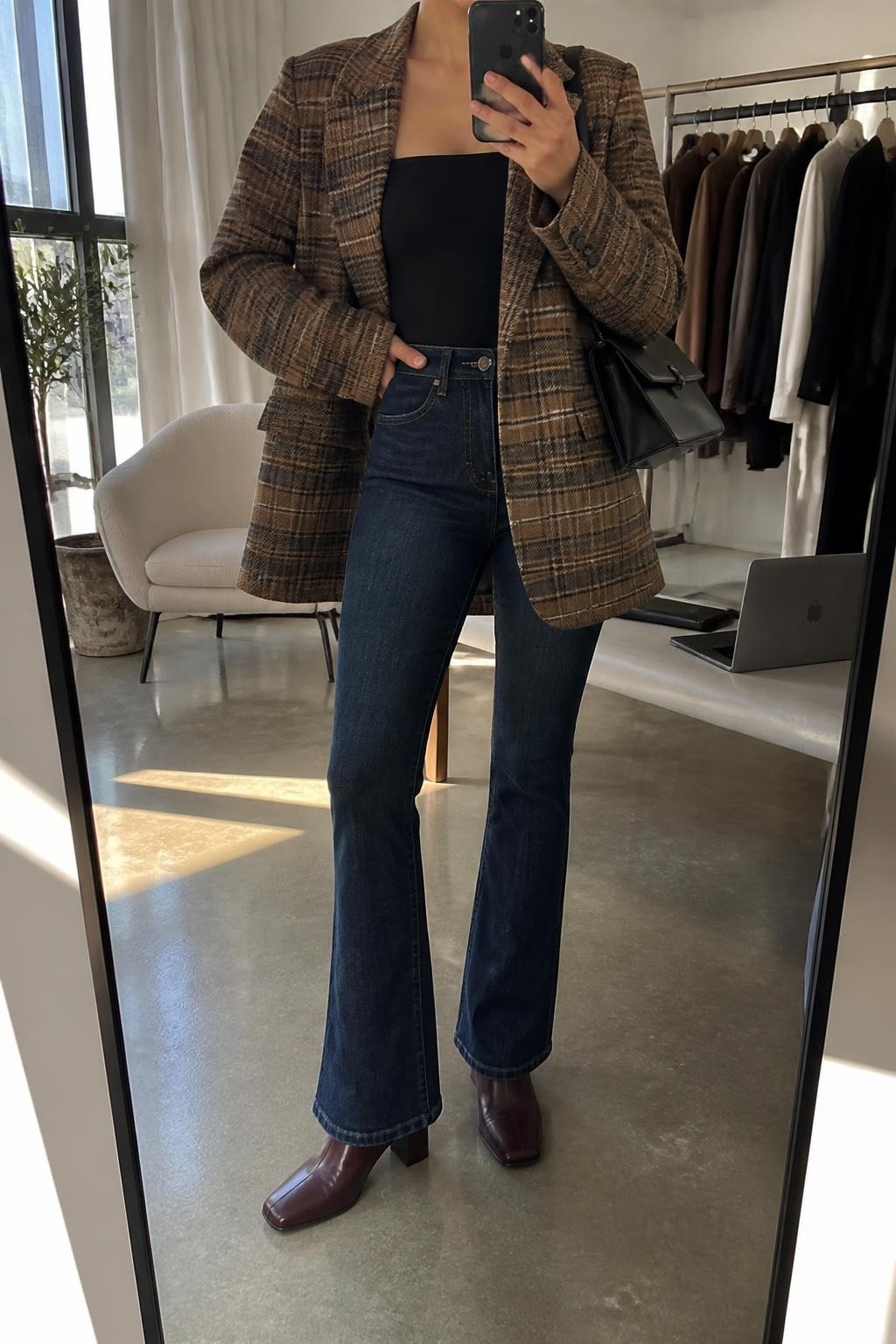 Mirror selfie of a woman wearing an oversized camel and charcoal plaid wool blazer, fitted black square-neck bodysuit, dark blue high-waisted flared jeans, burgundy leather block-heel boots, and a structured black shoulder bag, clearly showing the exact clothing items, colors, fabrics, and accessories listed, holding a phone in front of a fashion studio mirror corner, cropped at the neck so the face does not show, relaxed confident selfie pose, natural window light, a clothing rack with garments behind and a laptop on a bench, polished realistic environment, latest iPhone photo quality.