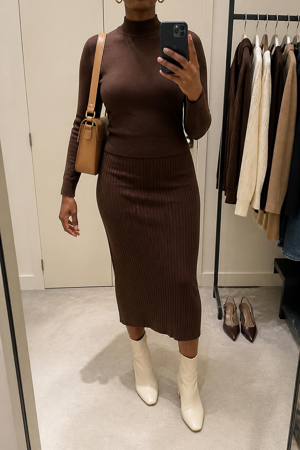 Mirror selfie of a Black woman wearing a chocolate-brown ribbed midi skirt, matching fitted chocolate mock-neck sweater, cream leather ankle boots, a tan structured shoulder bag, and small gold hoop earrings, clearly showing the exact clothing items, colors, fabrics, and accessories listed, holding a phone in front of a minimal dressing room mirror, cropped at the neck so the face does not show, relaxed casual selfie pose, soft indoor lighting, a clothing rack with garments behind and a pair of heels on the floor nearby, realistic and polished, latest iPhone photo quality.