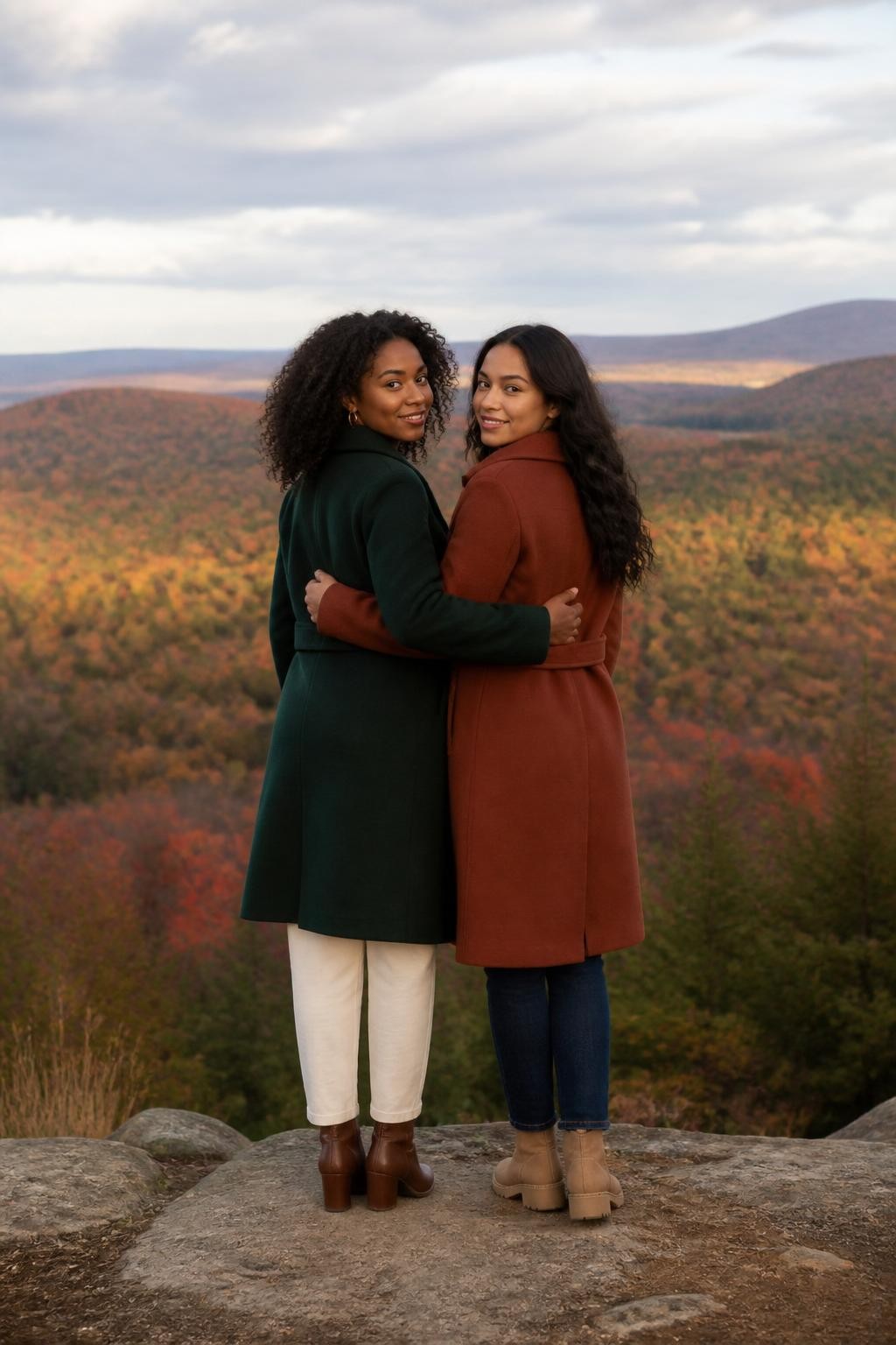 Image Prompt: Two mixed-race female best friends standing at a safe hilltop overlook during peak fall color, one Black woman wearing a deep green coat with cream trousers and brown boots, her friend wearing a rust coat, dark jeans, and tan boots, both standing in a loose side hug with arms around each other’s backs while looking toward the wide autumn valley, then turning slightly toward the camera with soft smiles, red, gold, and green trees stretching into the distance, gentle late-afternoon light beneath mild cloud cover, photographed with a DSLR 35mm lens from a slightly low angle, realistic landscape depth, natural skin detail, visible clothing folds, no unsafe cliff position, no dramatic artificial sun rays.