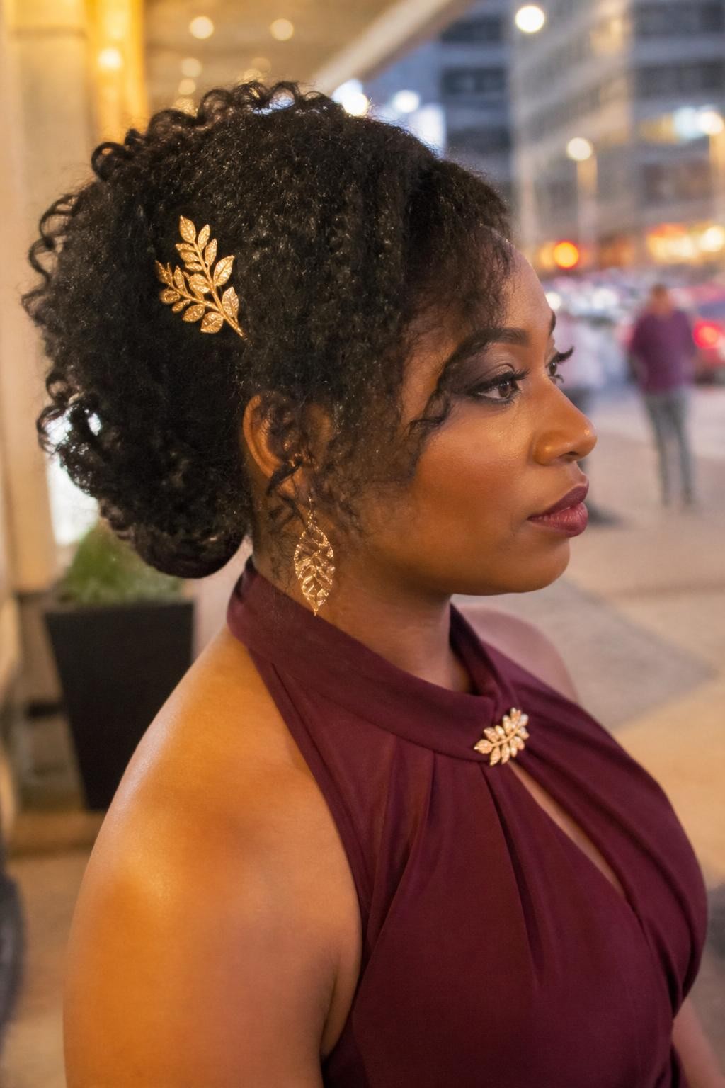 Image prompt: Photo of a Black woman wearing an elegant side-swept curly updo with textured curls gathered above one shoulder and a small gold leaf pin, side profile view, softly blurred city street background outside an evening venue, golden hour glow, containing statement earrings peeking through and a hair clip attached to her dress collar, realistic event moment, latest iPhone photo quality.