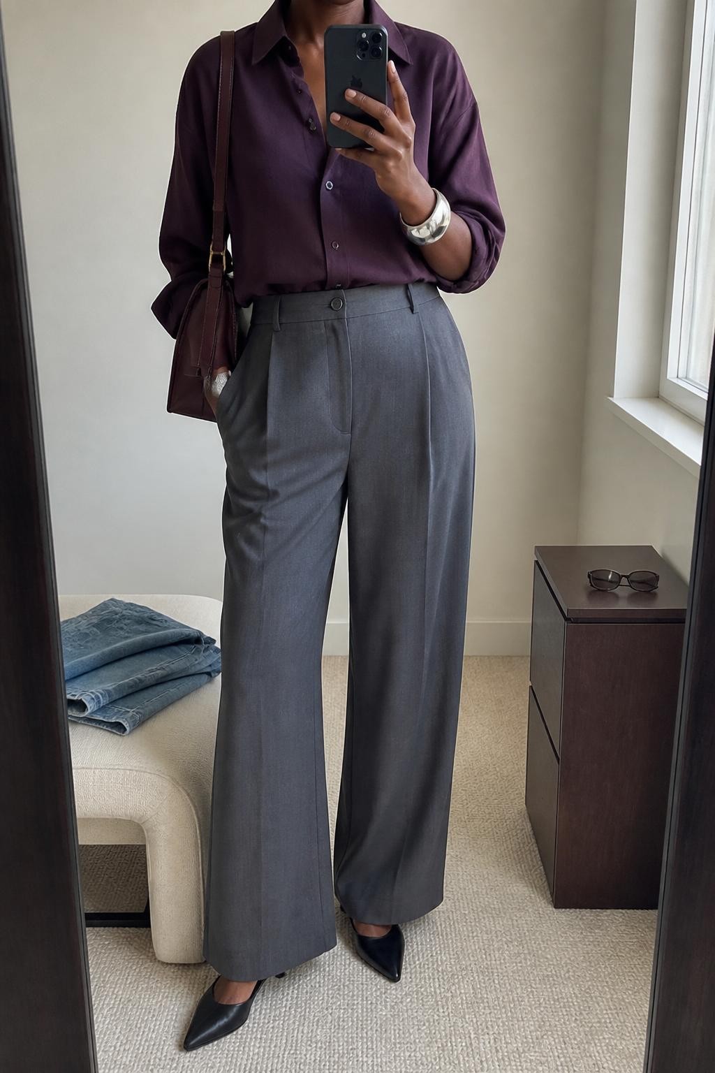 mirror selfie of a Black woman wearing the exact outfit described above, clearly showing a plum button-down shirt tucked into gray wide-leg pants, black pointed mules, a burgundy shoulder bag, and a silver cuff bracelet, holding a phone in front of a mirror, cropped at NECK so the face does not show, with a relaxed casual selfie pose, in a minimal dressing room, natural window light, with folded denim jeans on a bench and sunglasses placed on a dresser, latest iPhone photo quality.