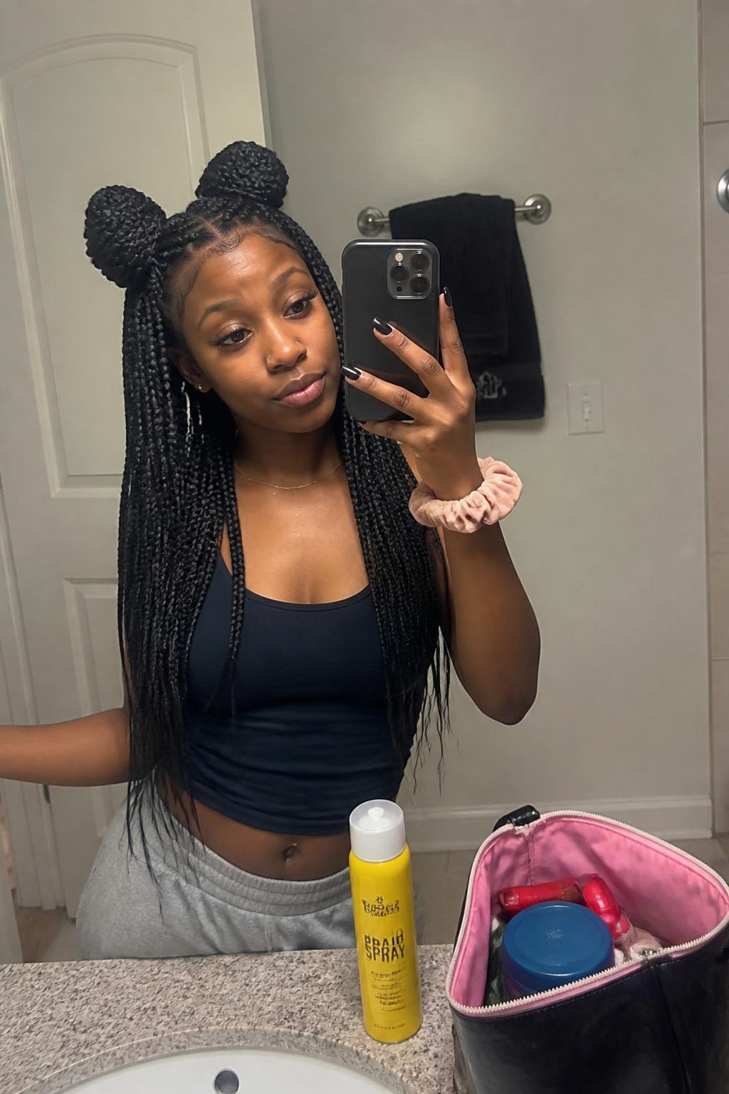 Image prompt: Photo of a young Black woman wearing long box braids styled into two half-up braided space buns with the remaining braids hanging loose, candid mirror selfie, bathroom vanity mirror with a makeup bag nearby, soft indoor lighting, containing a braid spray bottle on the counter and a scrunchie around her wrist, realistic casual styling moment, latest iPhone photo quality.