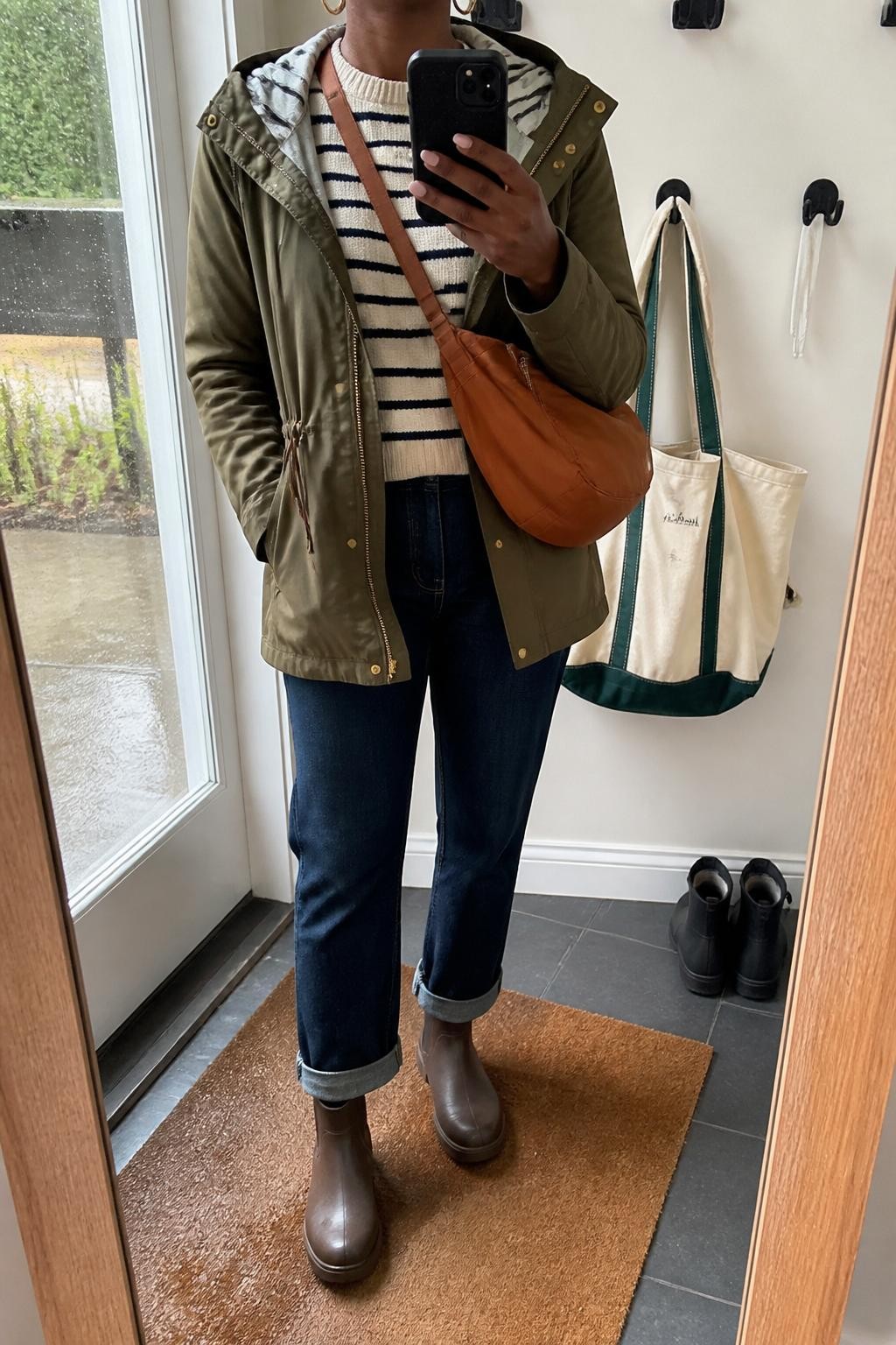 mirror selfie of a Black woman wearing the exact outfit described above, clearly showing an olive hooded anorak layered over a striped cream and navy sweater, dark blue straight-leg jeans cuffed above brown waterproof ankle boots, a cognac nylon crossbody bag, and small gold hoop earrings, holding a phone in front of a mirror, cropped at NECK so the face does not show, with a relaxed casual selfie pose, in a small mudroom-style entry mirror, natural rainy daylight, with a damp doormat near the boots and a tote bag hanging on a wall hook, latest iPhone photo quality.