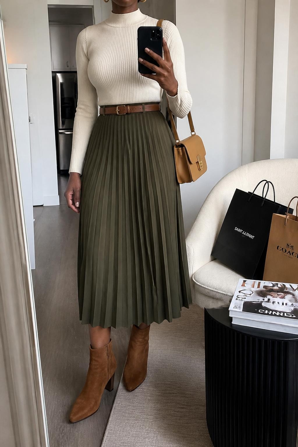 mirror selfie of a Black woman wearing the exact outfit described above, clearly showing cinnamon brown suede ankle boots, an olive pleated midi skirt, a fitted cream mock-neck sweater, a brown leather belt, a camel shoulder bag, and small gold hoop earrings, holding a phone in front of a mirror, cropped at NECK so the face does not show, with a relaxed casual selfie pose, in a stylish city apartment, soft indoor lighting, with shopping bags beside a chair and a stack of fashion magazines on a side table, latest iPhone photo quality.
