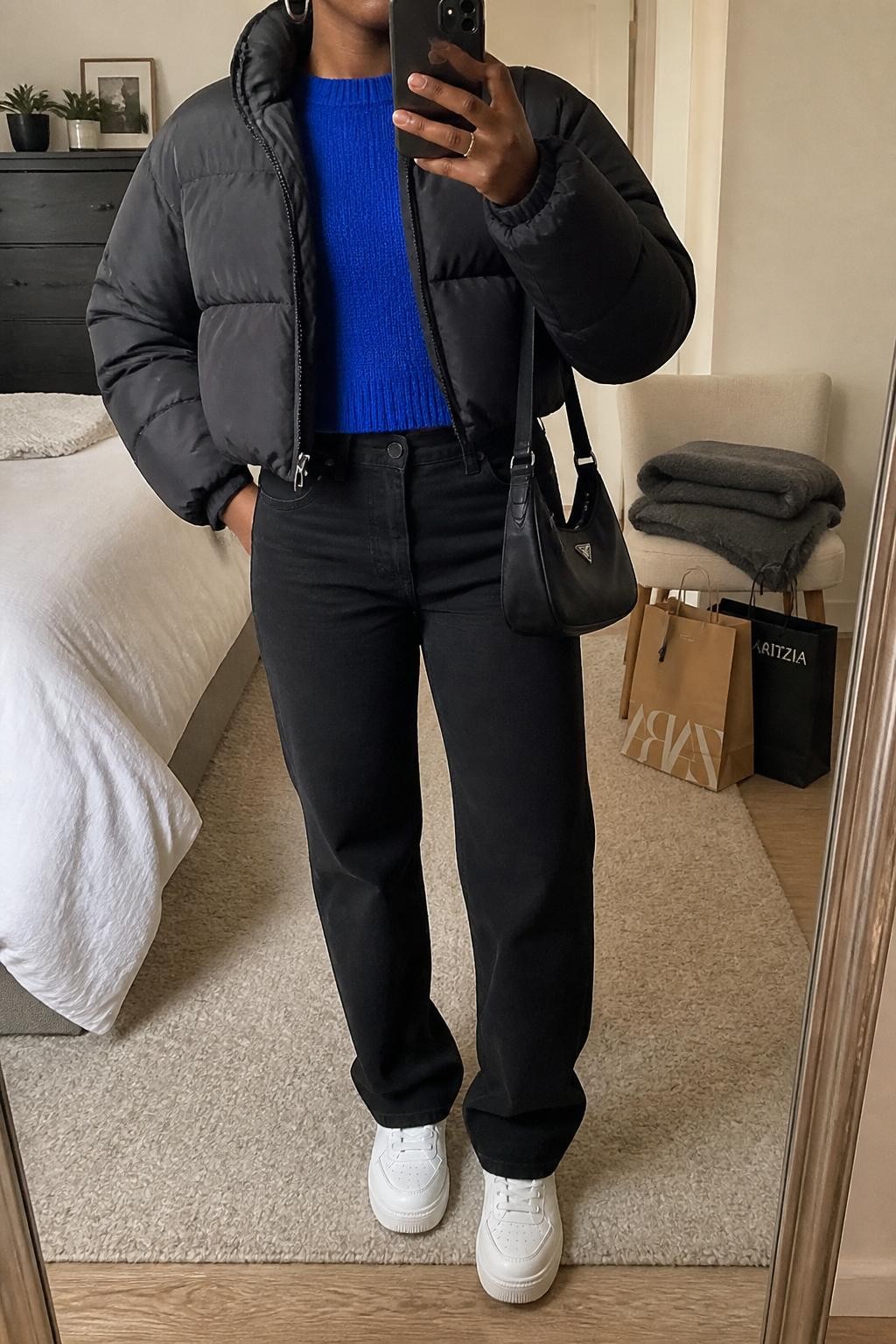 mirror selfie of a Black woman wearing the exact outfit described above, clearly showing a black cropped puffer jacket layered over an electric blue crewneck sweater, black straight-leg jeans, white platform sneakers, a black nylon shoulder bag, and silver hoop earrings, holding a phone in front of a mirror, cropped at NECK so the face does not show, with a relaxed casual selfie pose, in a cozy bedroom mirror setup, soft indoor lighting, with shopping bags beside a chair and a folded blanket on a chair, latest iPhone photo quality.