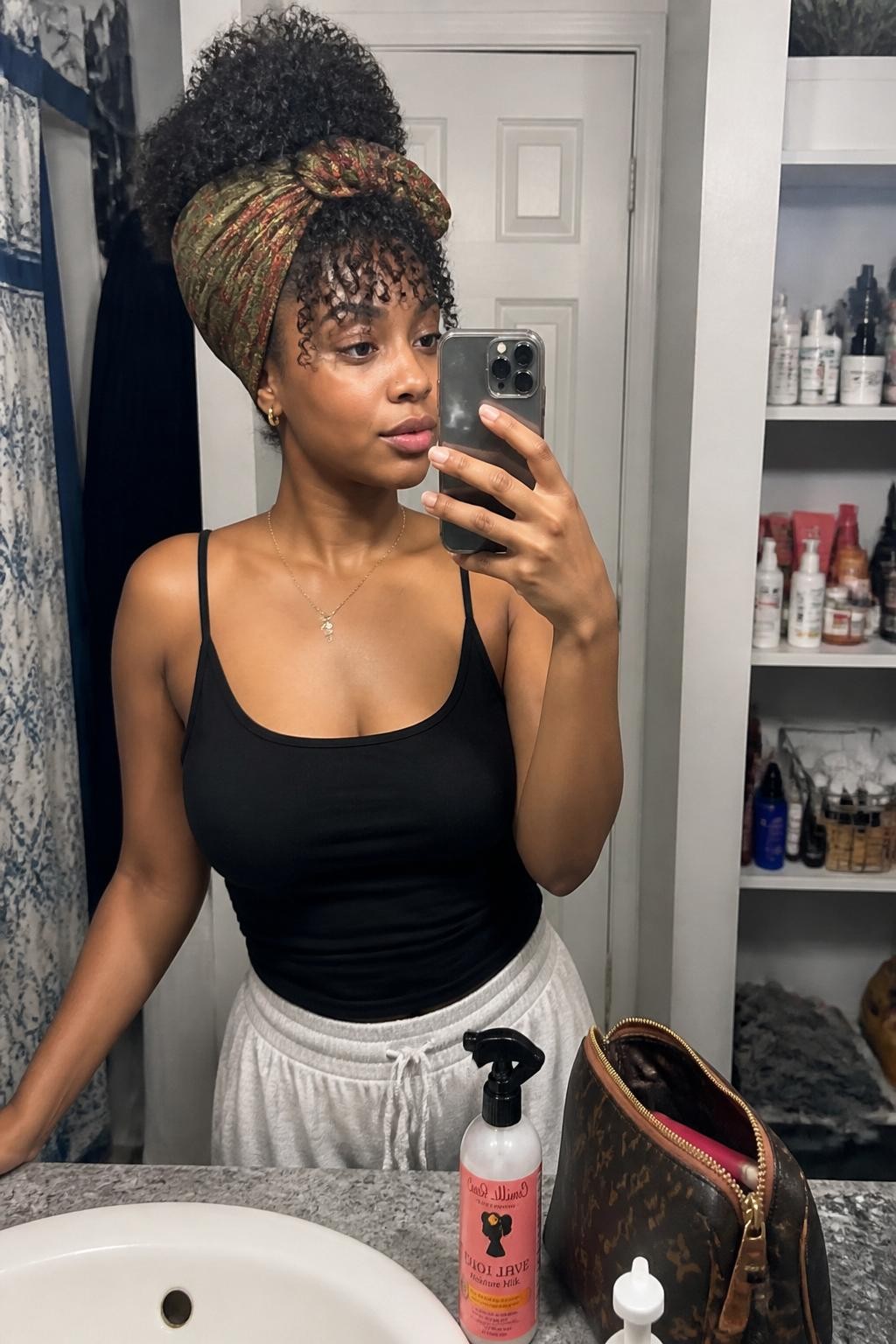 Image prompt: Photo of a Black woman wearing a high curly pineapple wrapped with an olive and rust printed scarf, candid mirror selfie, bathroom vanity mirror with skincare products nearby, soft indoor lighting, containing a curl spray bottle on the counter and a makeup bag beside the sink, realistic getting-ready moment, latest iPhone photo quality.