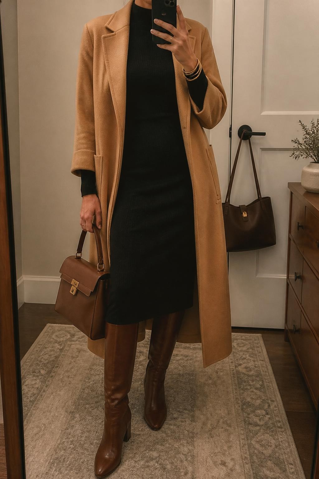 mirror selfie of a woman wearing the exact outfit described above, clearly showing whiskey brown leather knee-high boots, a black long-sleeve sweater midi dress, a camel scarf coat worn open, a whiskey brown top-handle bag, and gold bangles, holding a phone in front of a mirror, cropped at NECK so the face does not show, with a relaxed casual selfie pose, in a minimal dressing room, warm moody lighting, with a handbag hanging on a door handle and a small plant on a dresser, latest iPhone photo quality.