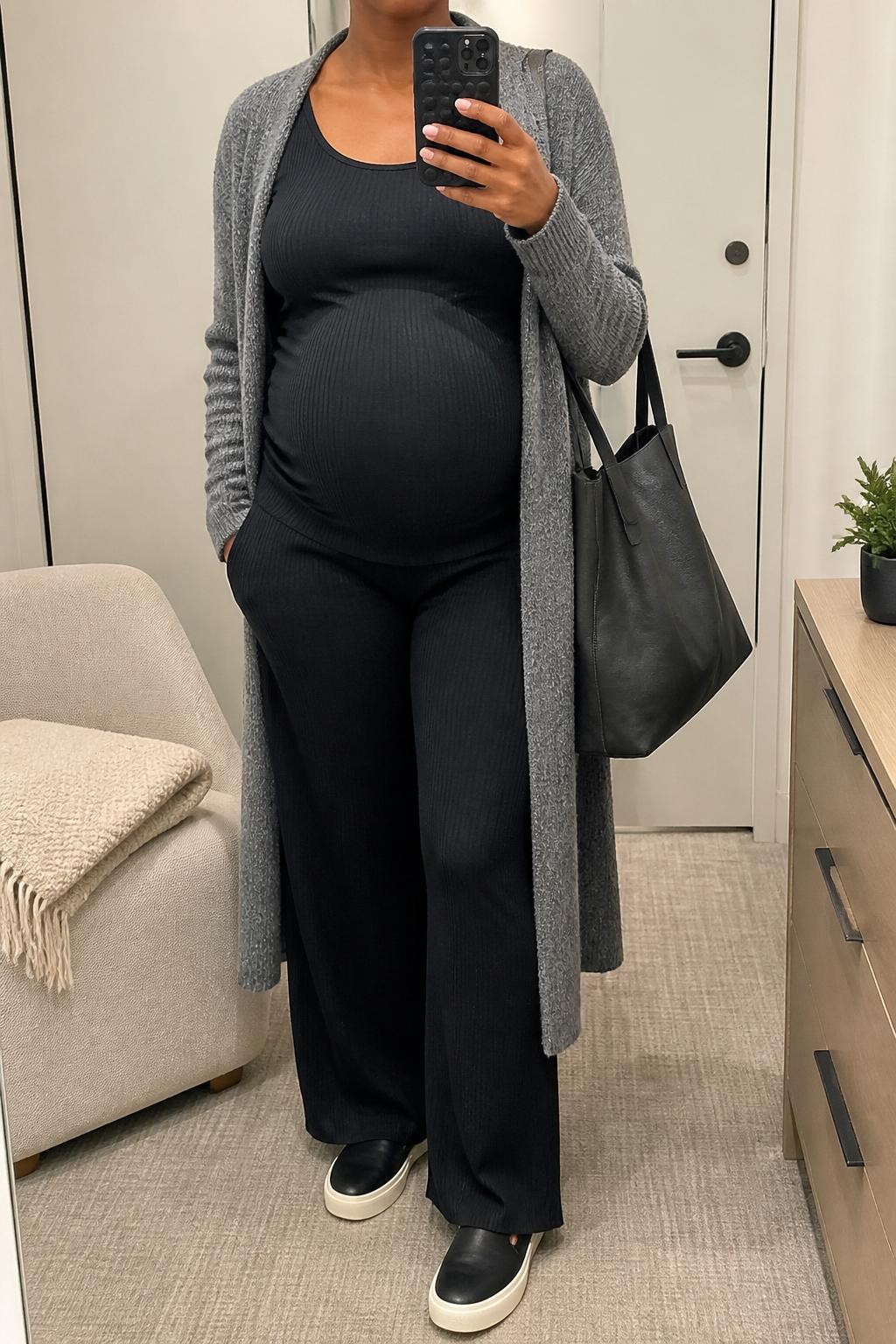 mirror selfie of a pregnant Black woman wearing a black ribbed maternity tank top, black ribbed maternity wide-leg pants, long gray sweater coat, black slip-on sneakers, and a charcoal tote bag, clearly showing the same clothing items, colors, fabrics, and accessories listed in the Outfit Pieces section, holding a phone in front of a mirror, cropped at NECK so the face does not show, with a relaxed casual selfie pose, in a minimal dressing room, soft indoor lighting, including folded blanket on a chair and a small plant on a dresser, latest iPhone photo quality.