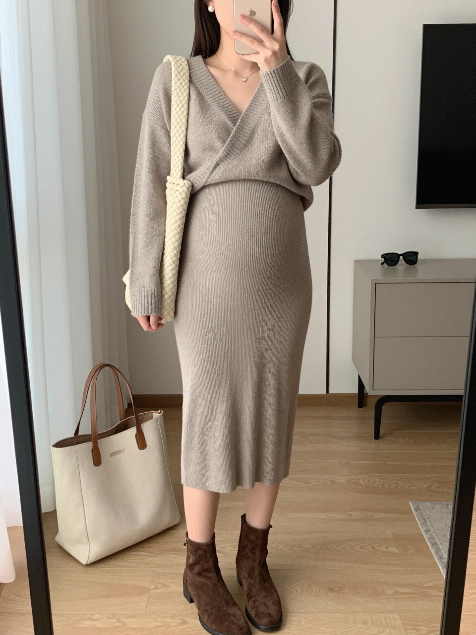 Mirror selfie of a pregnant woman wearing a fitted taupe ribbed maternity midi skirt, a matching taupe wrap-front knit sweater tied above the bump, chocolate-brown suede ankle boots, a cream woven shoulder bag, and small pearl stud earrings, clearly showing the exact clothing items, colors, fabrics, and accessories listed, holding a phone in front of a stylish city apartment mirror, cropped at the neck so the face does not show, with a relaxed casual selfie pose, soft indoor lighting, a tote bag resting on the floor and sunglasses placed on a dresser, natural and uncluttered, latest iPhone photo quality.