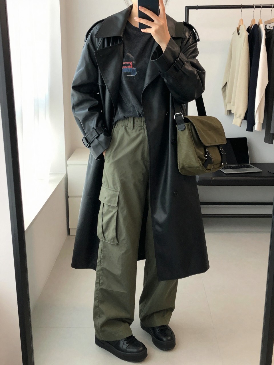 Mirror selfie of a woman wearing loose olive-green utility pants, a fitted faded black graphic T-shirt, a long black faux-leather trench coat, black platform sneakers, and a dark olive messenger bag, clearly showing the exact clothing items, colors, fabrics, and accessories listed, holding a phone in front of a minimal dressing room mirror, cropped at the neck so the face does not show, relaxed casual selfie pose, natural window light, a clothing rack with garments behind and a laptop on a bench, modern realistic setting, latest iPhone photo quality.