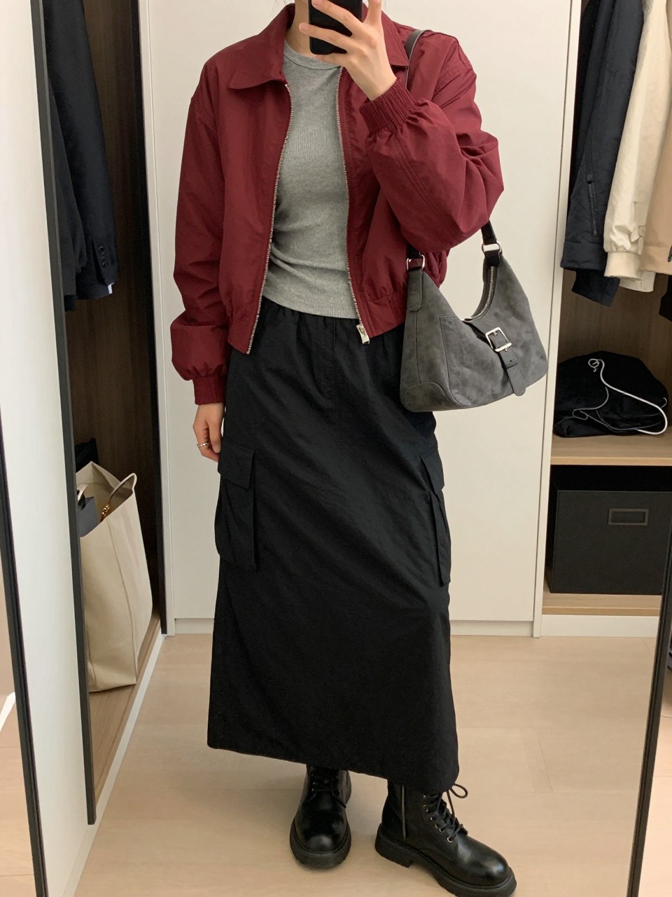 Mirror selfie of a woman wearing a black nylon cargo maxi skirt with side pockets, a fitted grey ribbed long-sleeve top, a cropped burgundy bomber jacket, black leather combat boots, and a charcoal-grey shoulder bag with silver buckles, clearly showing the exact clothing items, colors, fabrics, and accessories listed, holding a phone in front of a walk-in closet mirror, cropped at the neck so the face does not show, relaxed casual selfie pose, soft indoor lighting, an open closet with clothes visible and a tote bag resting on the floor, casual and realistic, latest iPhone photo quality.