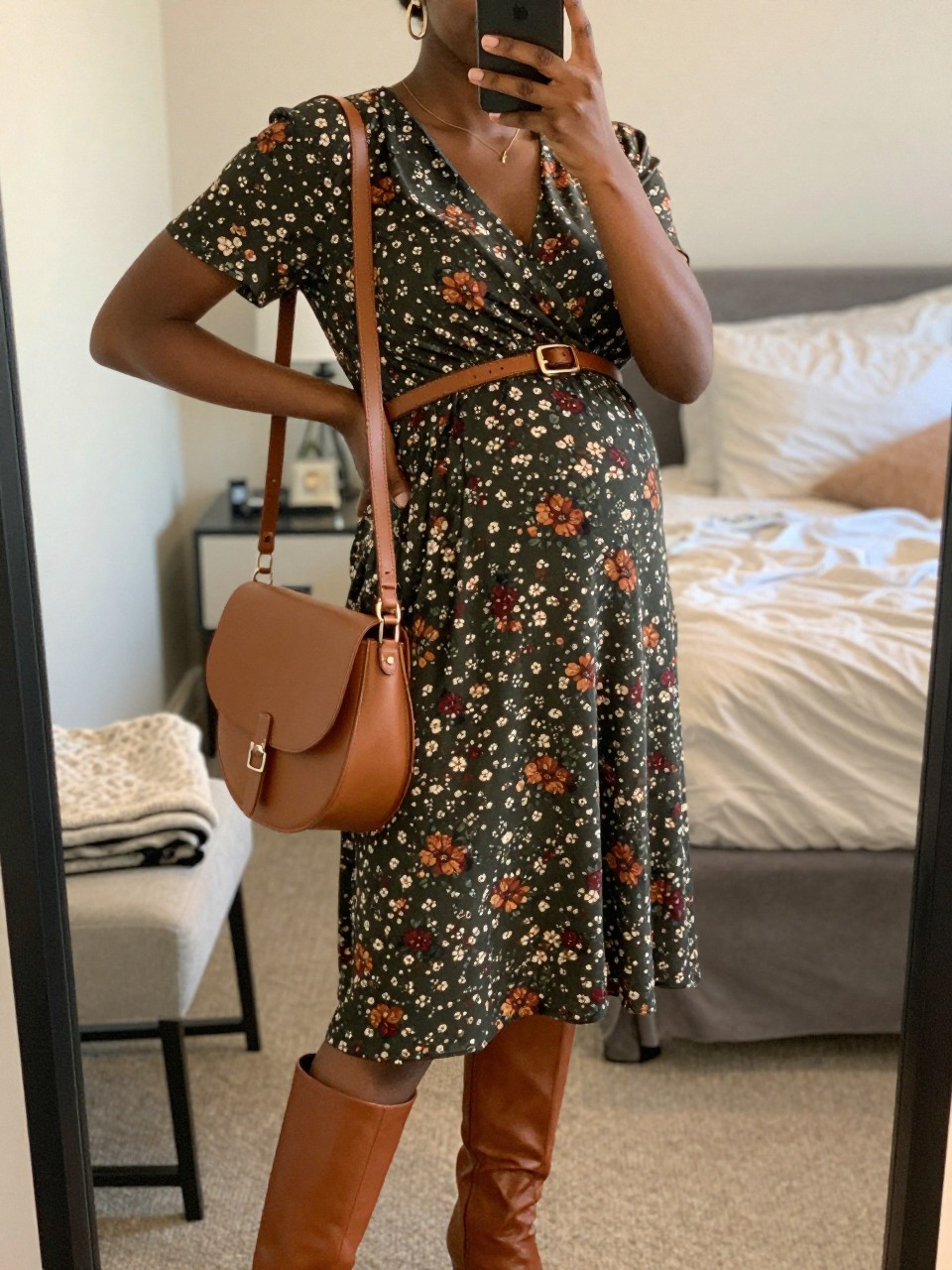 Mirror selfie of a Black pregnant woman wearing a dark olive maternity wrap midi dress with small rust, cream, and burgundy floral prints, cognac leather knee-high boots, a slim brown leather belt positioned above the bump, a tan saddle bag, and small hammered-gold earrings, clearly showing the exact clothing items, colors, fabrics, and accessories listed, holding a phone in front of a cozy bedroom mirror setup, cropped at the neck so the face does not show, with a relaxed casual selfie pose, soft indoor lighting, soft pillows on an unmade bed and a folded blanket on a chair, believable lived-in setting, latest iPhone photo quality.
