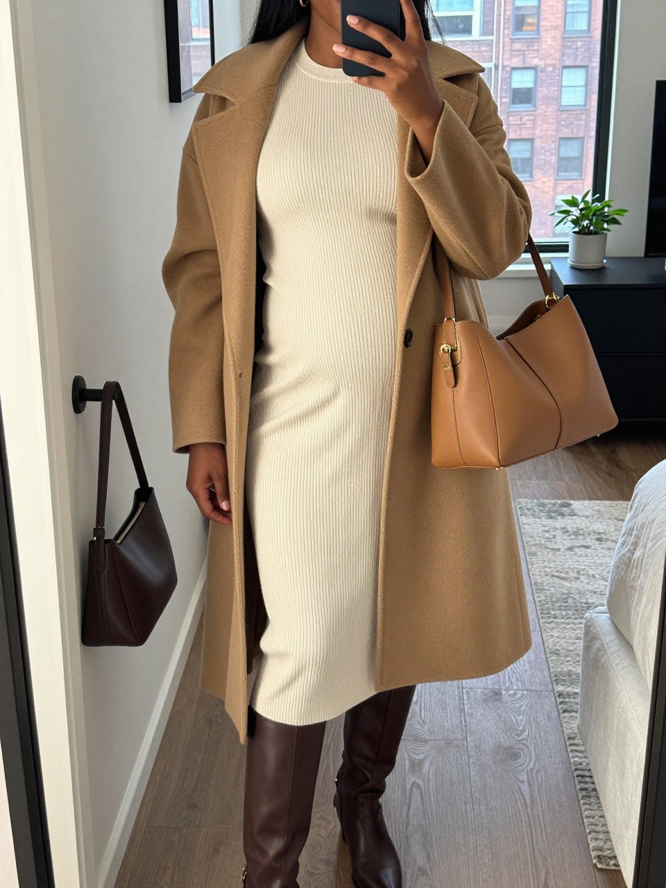 Mirror selfie of a Black pregnant woman wearing a fitted cream ribbed-knit maternity midi dress, an open camel wool wrap coat, chocolate-brown knee-high leather boots, a tan structured handbag, and small gold hoop earrings, clearly showing the exact clothing items, colors, fabrics, and accessories listed, holding a phone in front of a stylish city apartment mirror, cropped at the neck so the face does not show, with a relaxed casual selfie pose, natural window light, a handbag hanging on a door handle and a small plant on a dresser, realistic and uncluttered, latest iPhone photo quality.